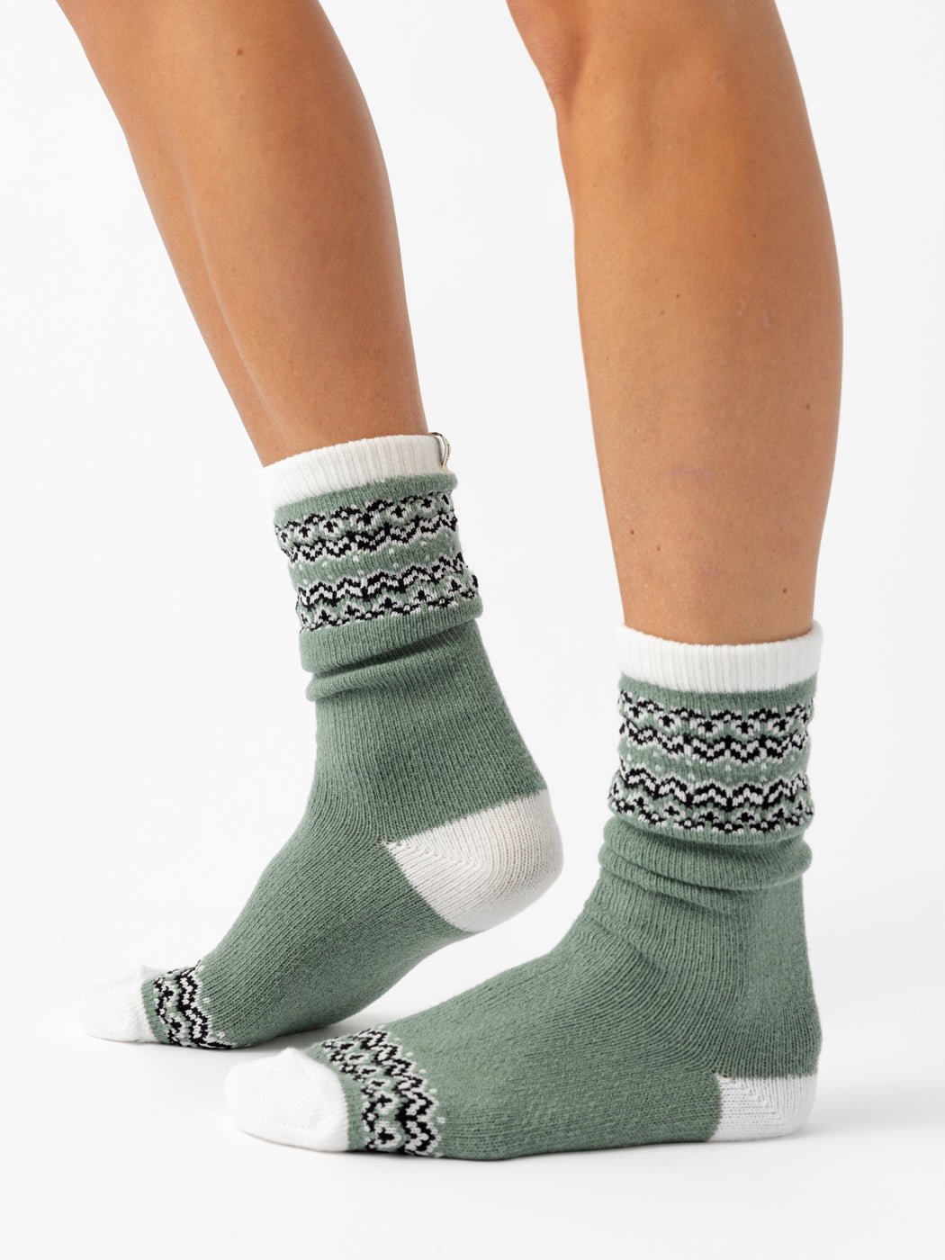 A person wears Cozy Earth’s The Fair Isle Plush Lounge Sock, a light green style with white toes, heels, and cuffs, plus black-and-white geometric patterns on the cuffs and near the toes. The background is white.