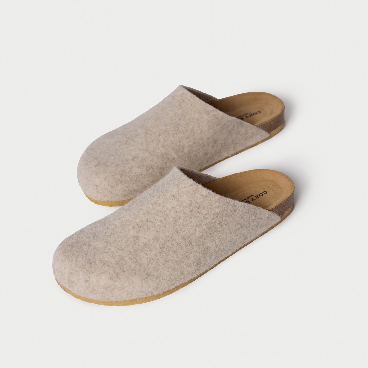 The Lakehouse Clog by Cozy Earth are beige woolen slip-on slippers with a cork sole, viewed from above. They feature a minimalist design perfect for indoor use against a light cream background. |Color:Oat