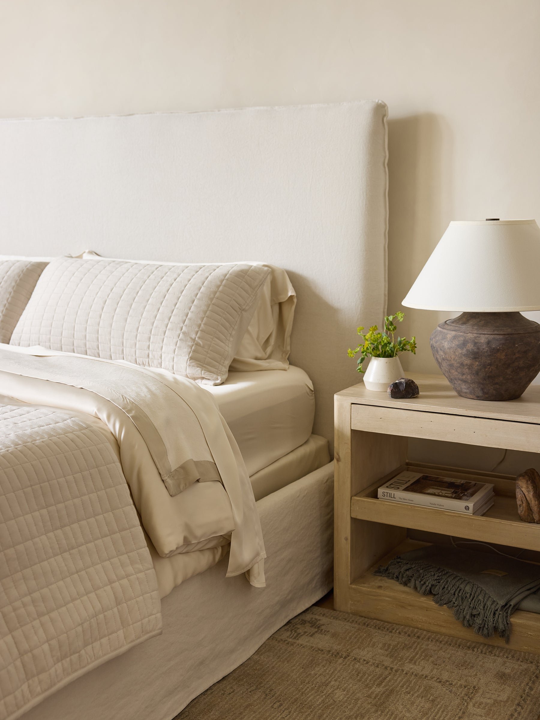 A cozy bedroom features a neatly made bed accentuated by Cozy Earth's Bamboo Blanket, boasting cream-colored bedding and a padded headboard. Beside the bed sits a wooden nightstand adorned with a ceramic table lamp, a small plant, and several books. Soft, neutral tones create an invitingly tranquil atmosphere. |Color:Oat/Driftwood