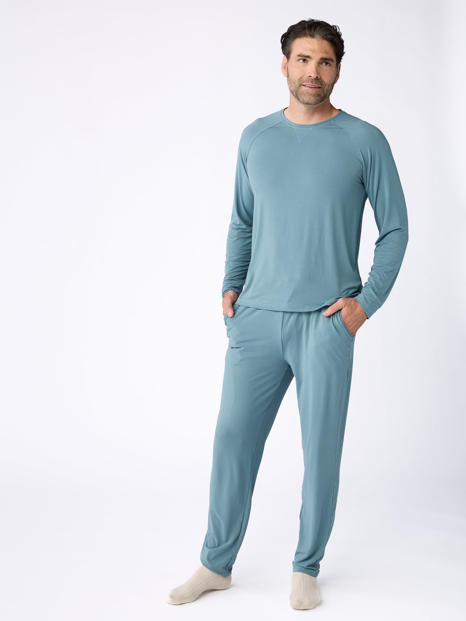 A person is dressed in a coordinated light blue loungewear set, featuring the Men's Stretch-Knit Bamboo Long Sleeve from Cozy Earth and matching pants. They are positioned against a plain white background, with their left hand resting in their pocket and sporting cream-colored socks. 