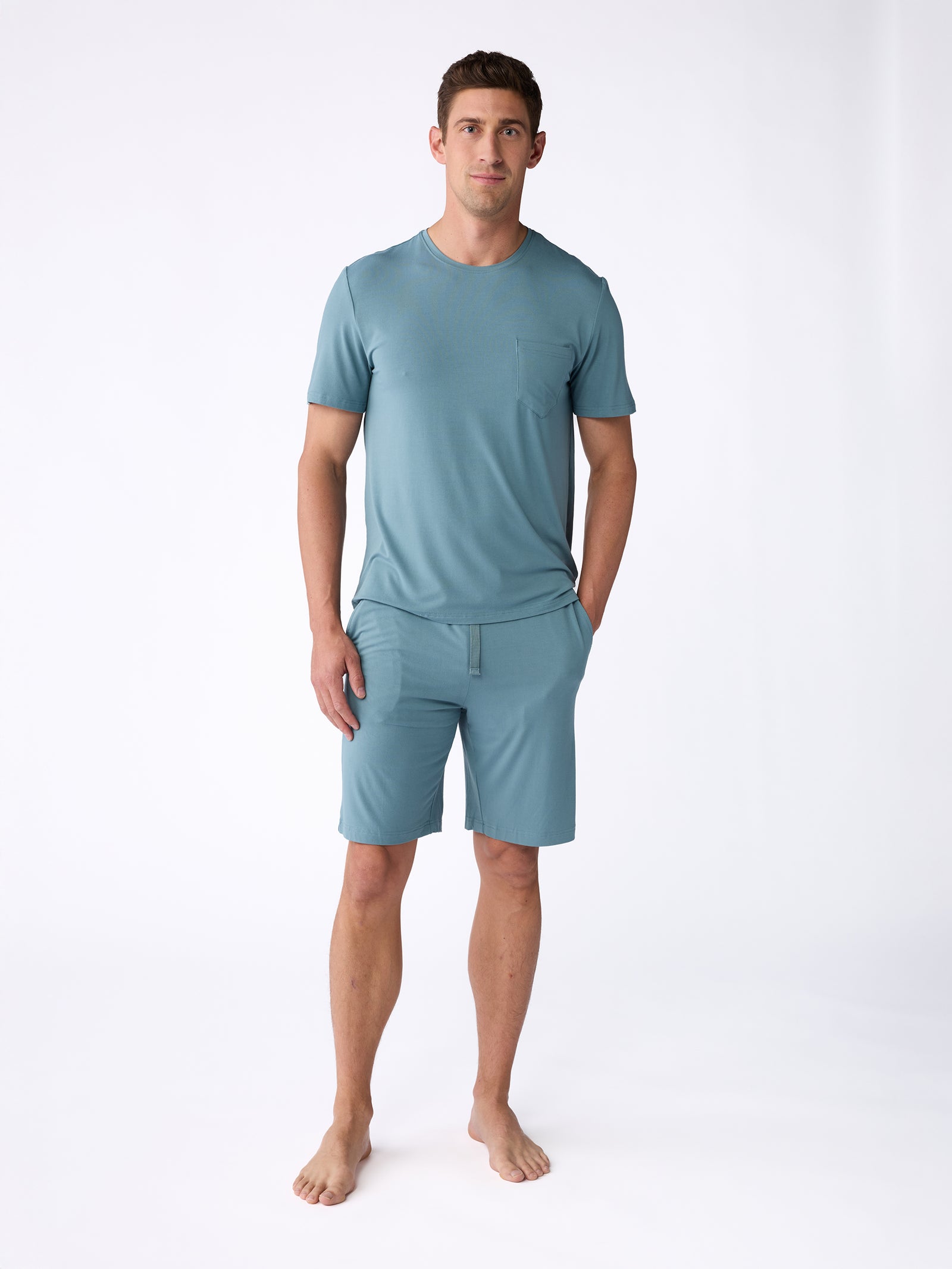 A man wearing the Men's Stretch-Knit Bamboo Pajama Short from Cozy Earth stands barefoot against a plain white background. His hands rest by his sides, and he gazes directly at the camera. 