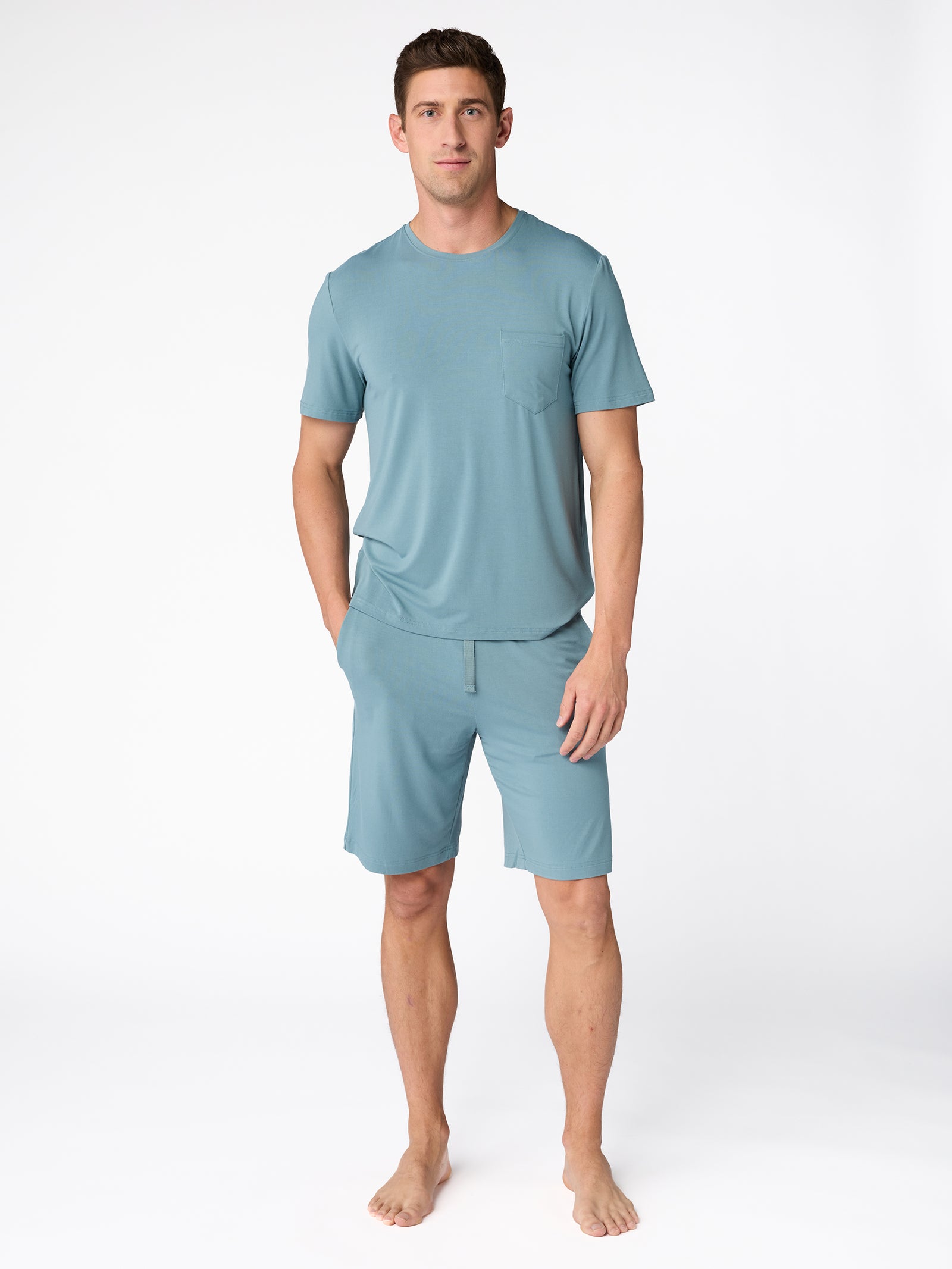 A man stands barefoot against a white background, wearing the Cozy Earth Men's Stretch-Knit Bamboo Lounge Tee in teal, along with matching shorts. One hand is in his pocket, and he has a relaxed expression. 