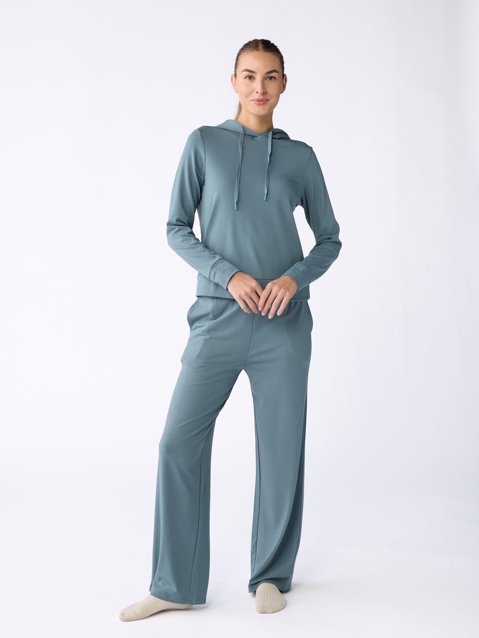 A person is seen wearing the Women's Ultra-Soft Bamboo Hoodie in teal from Cozy Earth. They are standing against a plain white background, smiling with their hands clasped in front, and beige socks complement their relaxed fit outfit. 