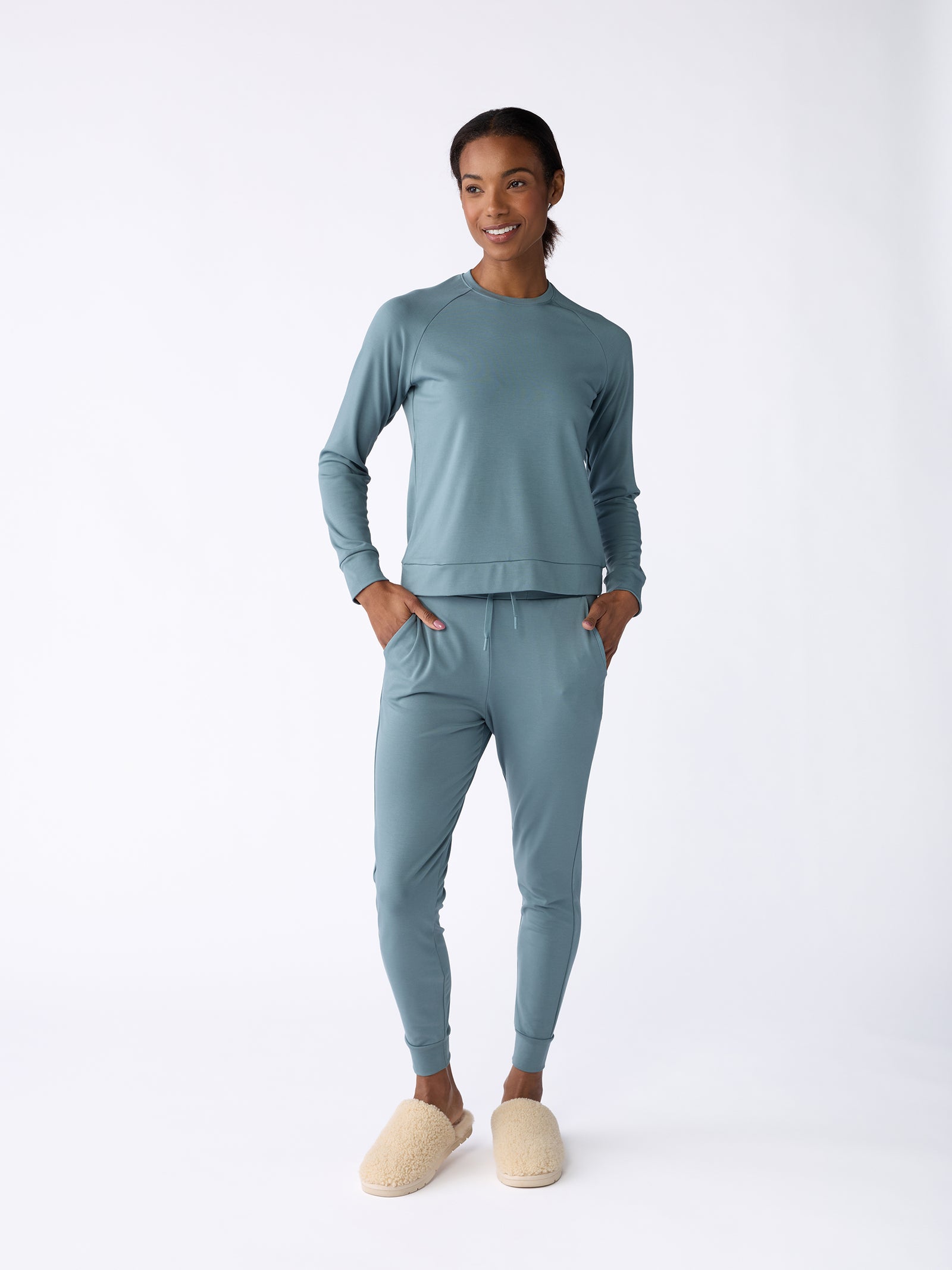 A person in a matching light blue ensemble, including the Women's Bamboo Jogger Pant from Cozy Earth, stands against a plain white background. They accessorize with cream-colored slippers and appear relaxed, with their hands in their pockets. 