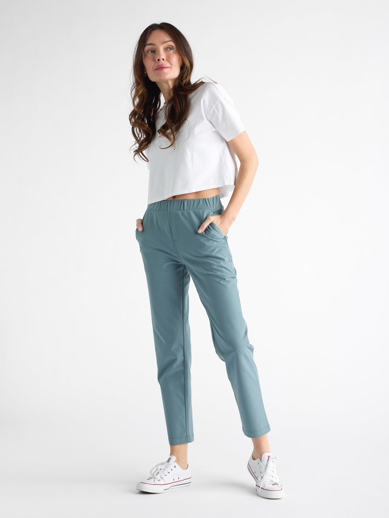 A person with long hair stands with hands in pockets, wearing a white cropped T-shirt, Cozy Earth's Women's Always Cropped Pant in teal, and white sneakers against a plain white background. 