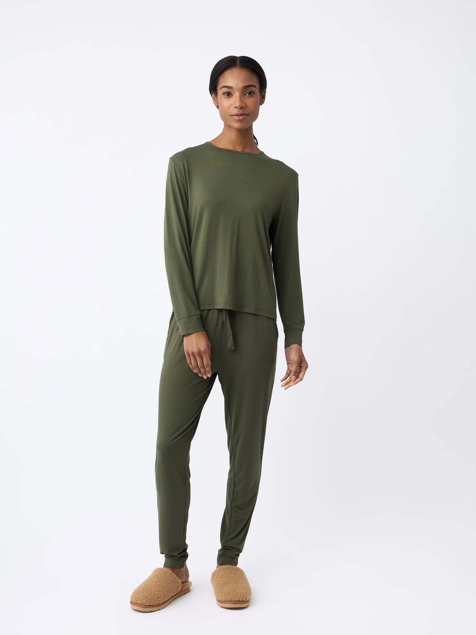 Against a plain white background, a woman wears the Women's Stretch Knit Bamboo Long Sleeve Lounge Tee in olive-green by Cozy Earth. They pair it with matching pants and fuzzy brown slippers. 