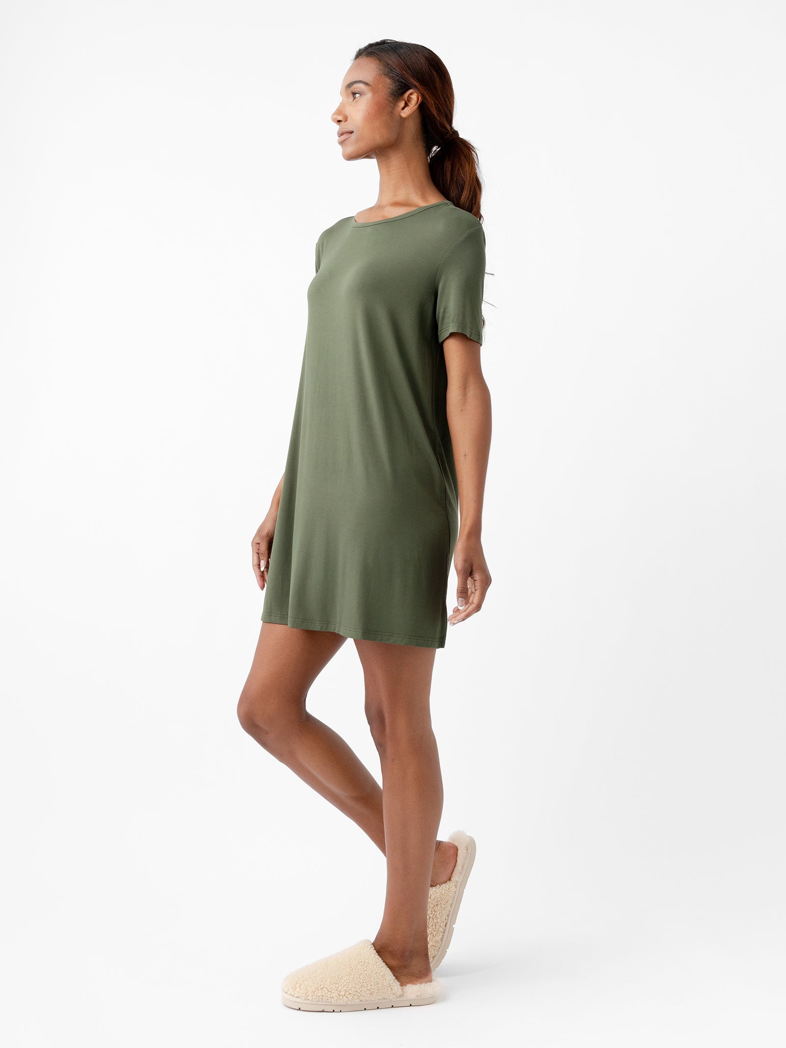 A person stands against a plain white background, wearing a Cozy Earth Women's Bamboo Stretch Knit Sleep Dress. The olive-green dress is loose-fitting, short-sleeved, and falls above the knees. They are also wearing beige slip-on shoes. The person gazes off to the side with a relaxed posture, one leg slightly bent. 