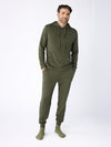 A man stands against a plain background wearing a matching green Men's Ultra-Soft Bamboo Hoodie and sweatpants from Cozy Earth, along with green socks. His hands are in his pockets, and he has a slight smile. |Color:Olive