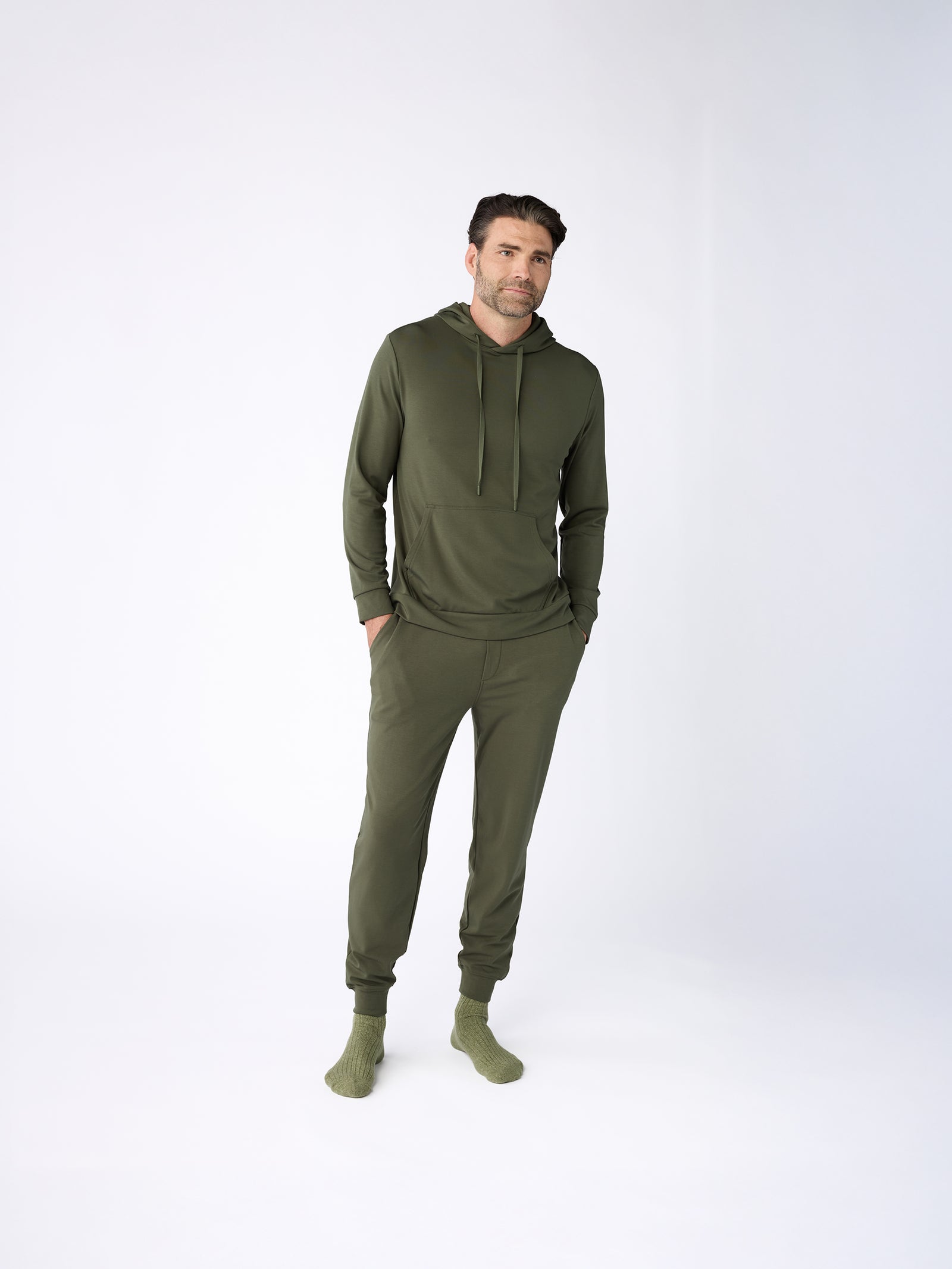 A man stands against a plain background wearing a matching green Men's Ultra-Soft Bamboo Hoodie and sweatpants from Cozy Earth, along with green socks. His hands are in his pockets, and he has a slight smile. 