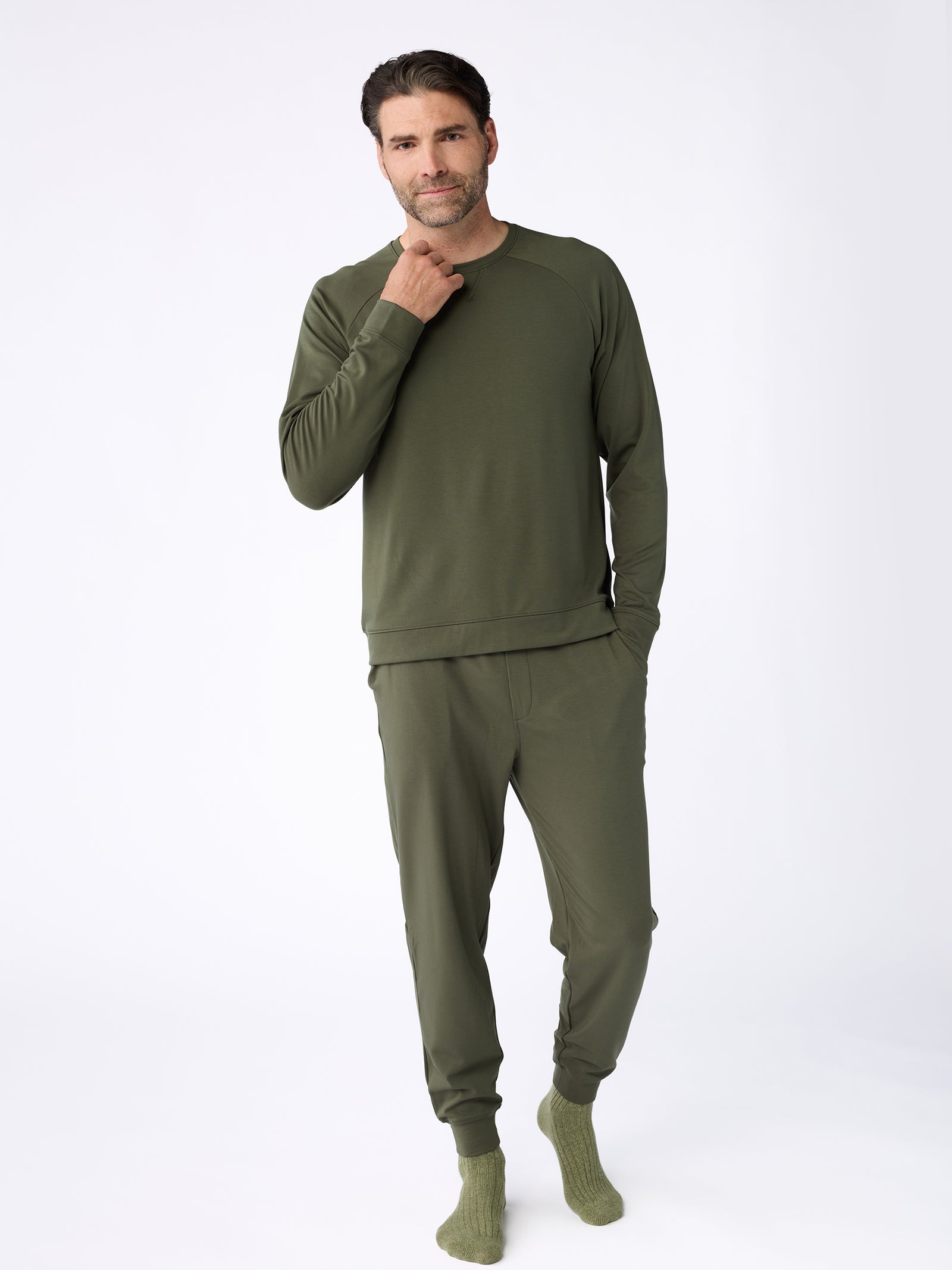 A man stands in a relaxed pose, wearing a matching olive green loungewear set from Cozy Earth, featuring the Men's Ultra-Soft Bamboo Pullover Crew sweatshirt and jogger pants. He has his right hand near his chin against a plain white background. 