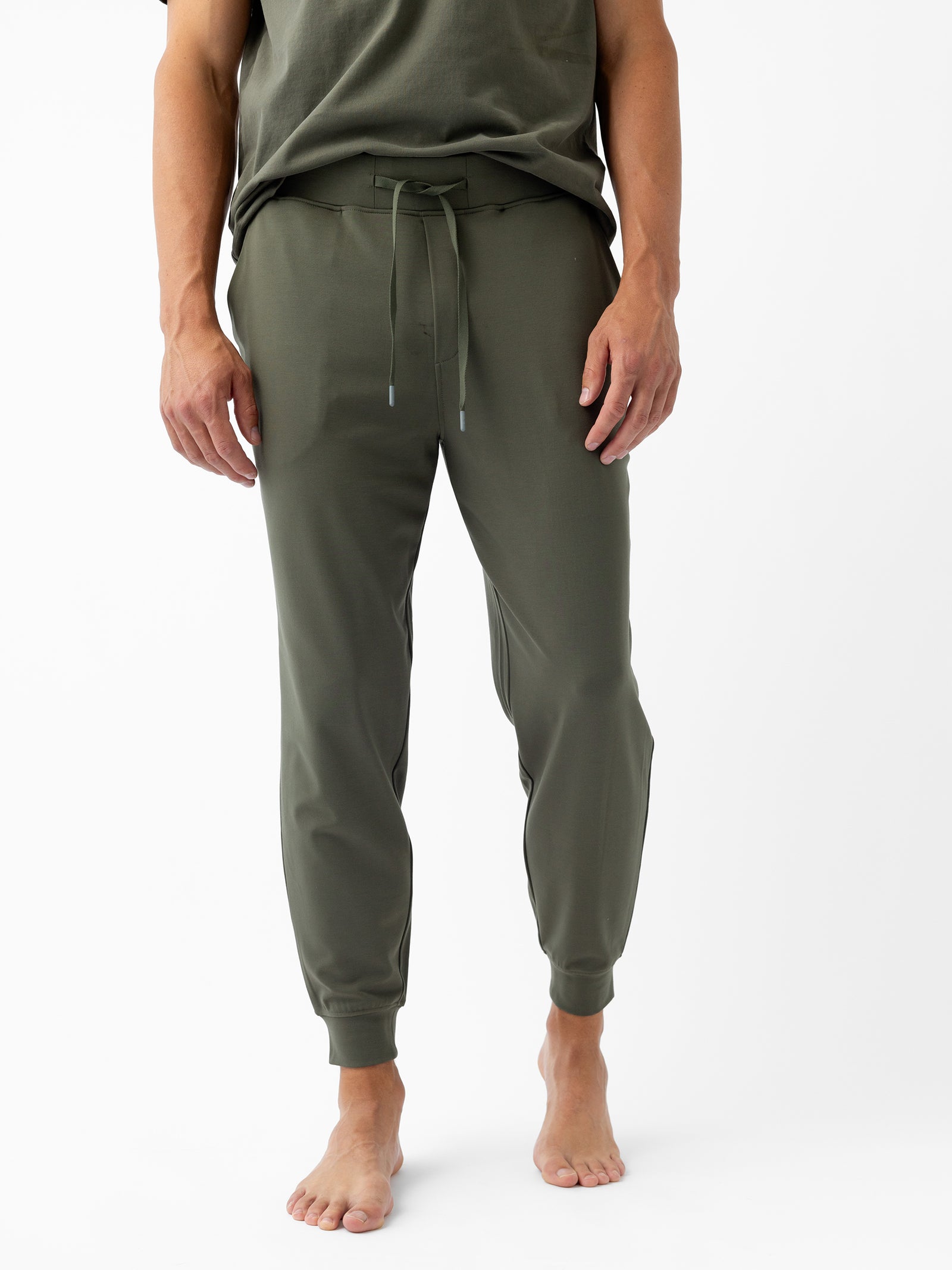 A person is standing against a white background wearing the Men's Ultra-Soft Bamboo Jogger Pant in olive green from Cozy Earth. The person is barefoot, with their hands by their sides, and a matching short-sleeved top is partially visible. 