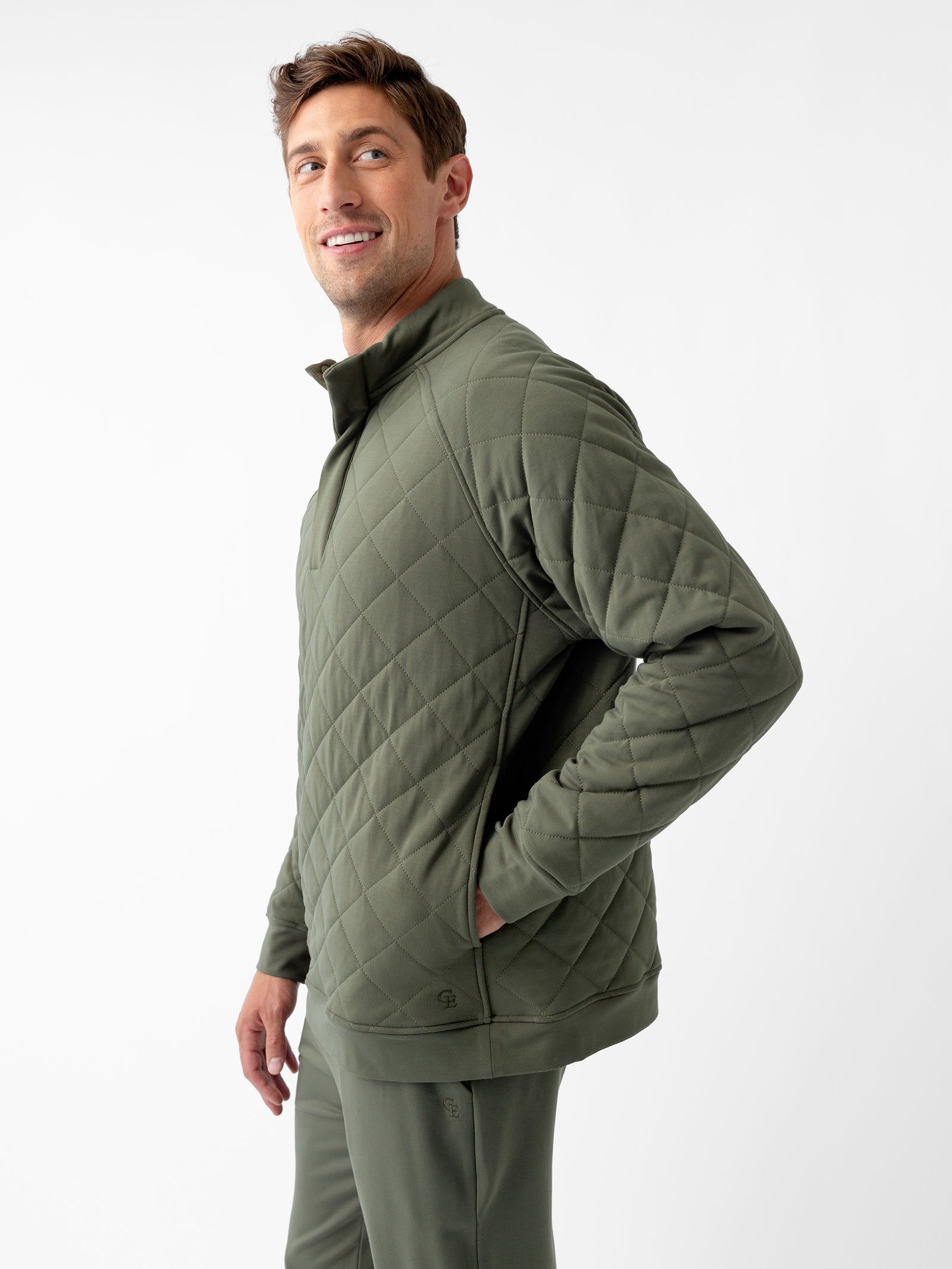 A person with short hair, wearing a Cozy Earth Men’s Ultra-Soft Bamboo Quilted Snap Pullover in olive green and matching pants, stands against a plain white background. The individual is smiling, has one hand in their pocket, and is slightly turning to the side. 