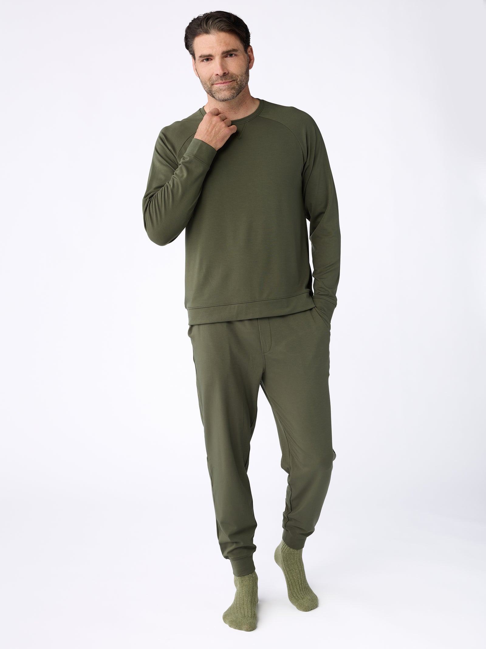 A man wearing the Men's Ultra-Soft Bamboo Pullover Crew from Cozy Earth, along with matching green pants, stands against a plain white background. He has one hand in his pocket and the other touching his chin, complemented by green socks. 