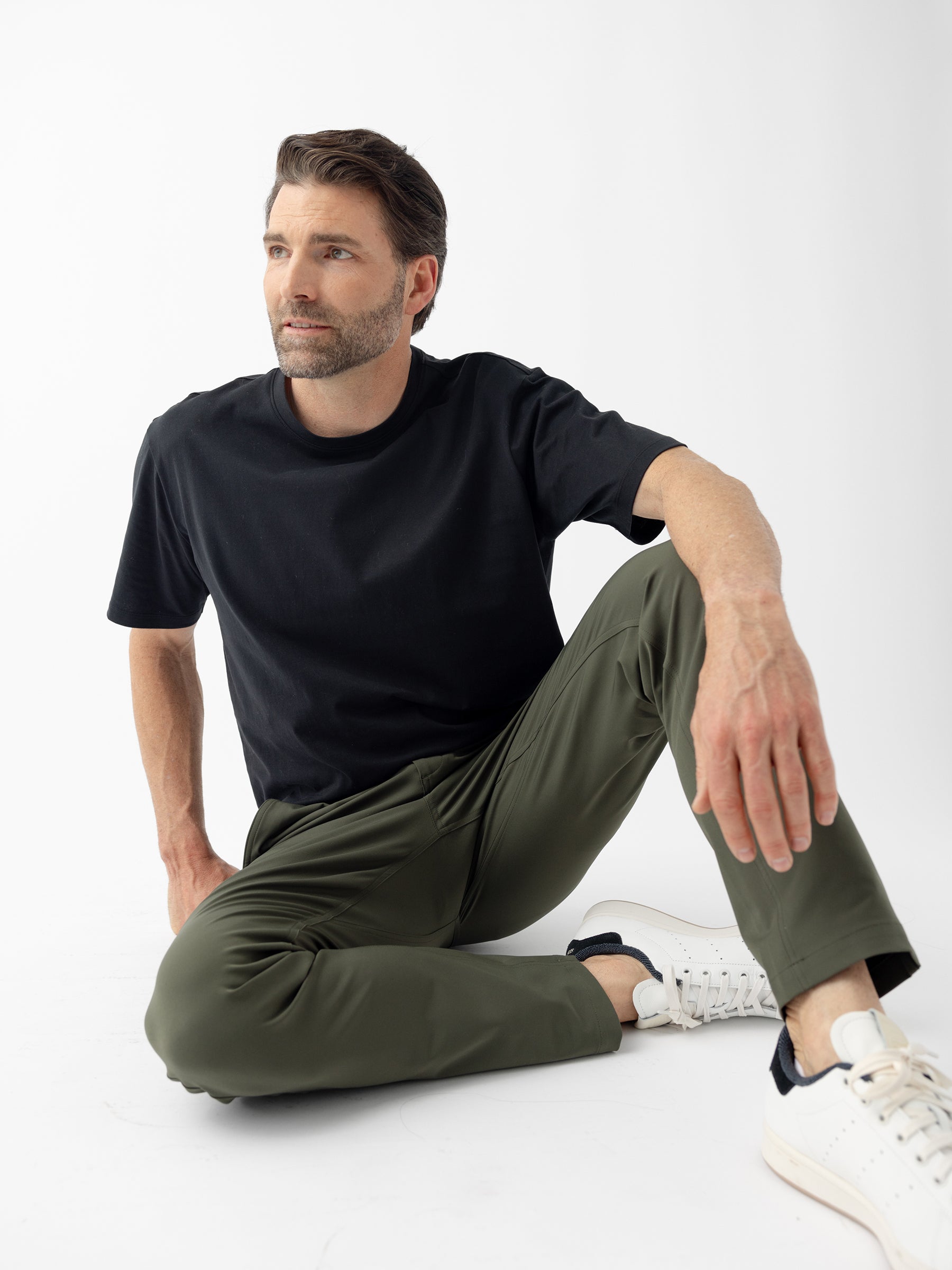 A man with short dark hair and a beard is seated on the ground against a plain white background. He is wearing a black T-shirt, Cozy Earth's olive green Men's Everywhere Pant 32L, and white sneakers with black detailing. He gazes off to the side, with one knee bent and the other leg folded under him. |Color:Olive