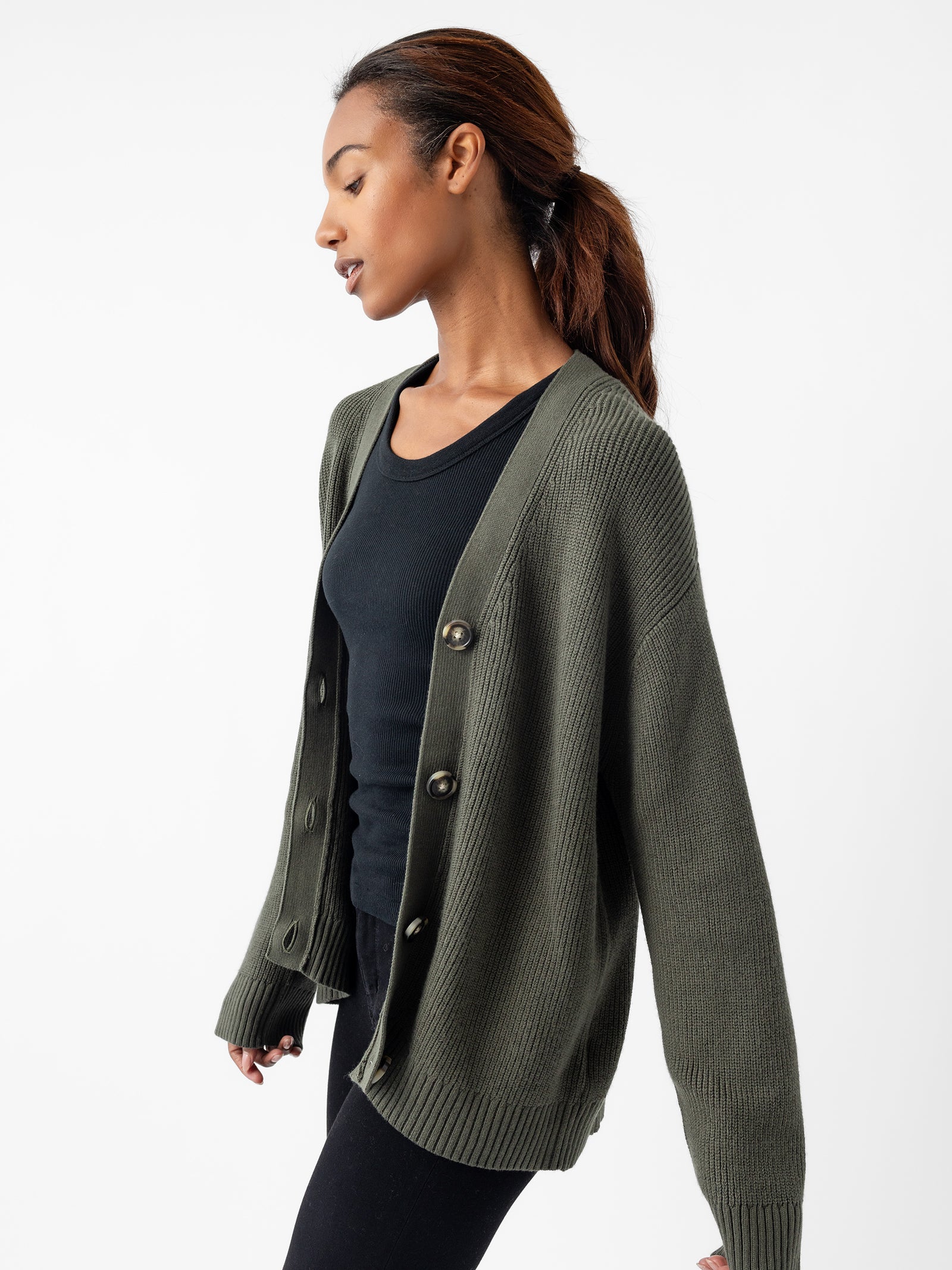 A person with long dark hair is wearing a black shirt and the Oversized Classic Cardigan by Cozy Earth in olive green, featuring button details. They are looking down and slightly turning to the side against a plain white background. 