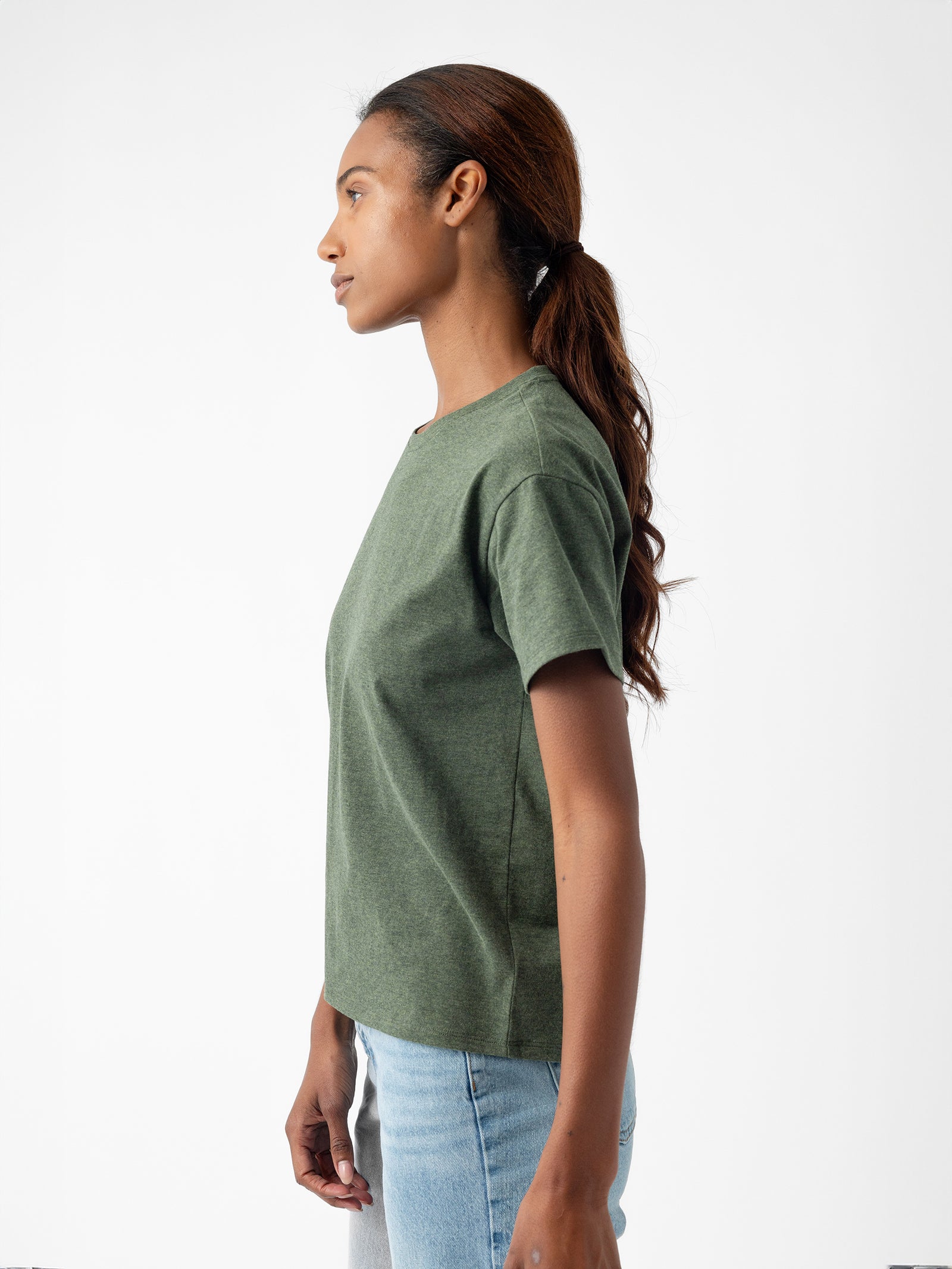 A woman with long, dark hair tied in a ponytail is standing in front of a plain white background, facing left. She is wearing Cozy Earth’s Women's All Day Tee in green and light blue jeans. Her arms are relaxed by her sides. 