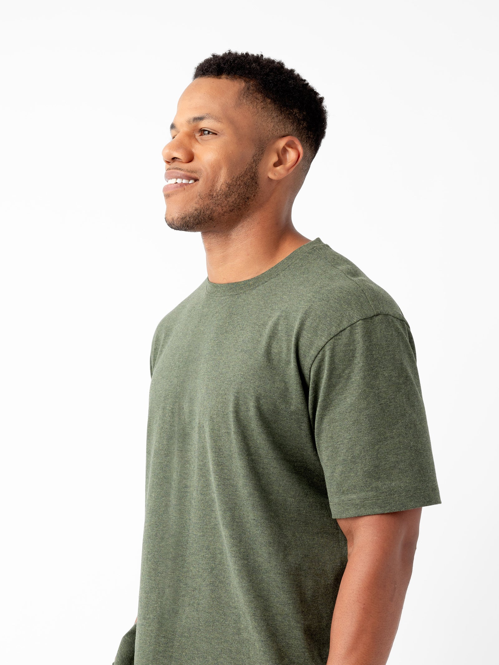 A person with short curly hair and a beard is smiling while looking to the side. They are wearing Cozy Earth's Men's All Day Tee in plain olive green and standing against a plain white background. 