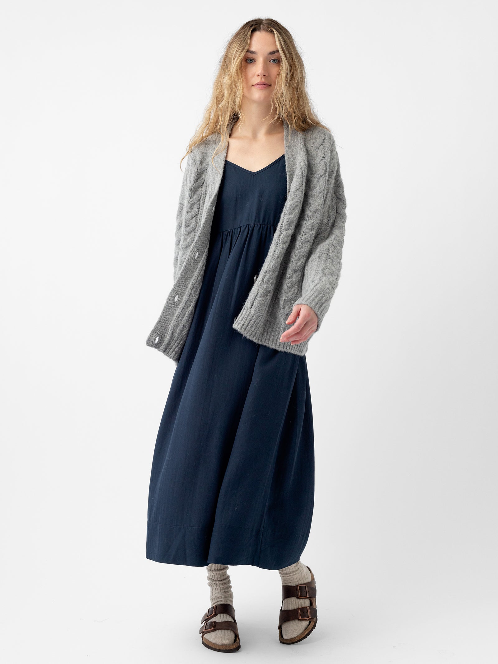 A person with long hair is standing against a plain white background, wearing a dark blue dress, an Oversized Cable Knit Cardigan by Cozy Earth in light gray, wool socks, and sandals. The outfit is casual and comfortable. 