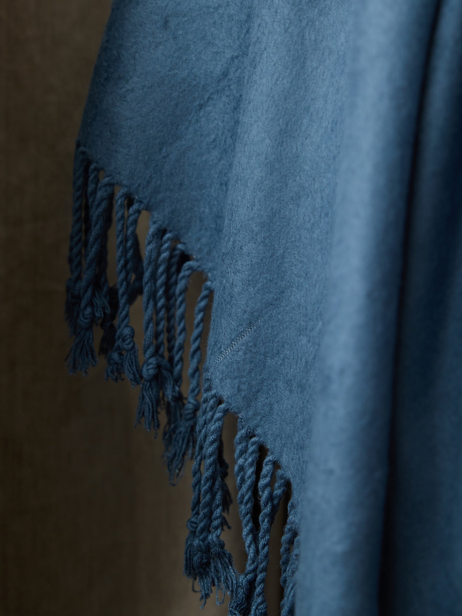 A close-up image of the Bamboo Tassel Throw by Cozy Earth in a soft blue hue. The fabric, appearing plush and fuzzy, is draped against a neutral background. Fringed tassel edges hang loosely, adding texture and detail to this luxurious throw. 