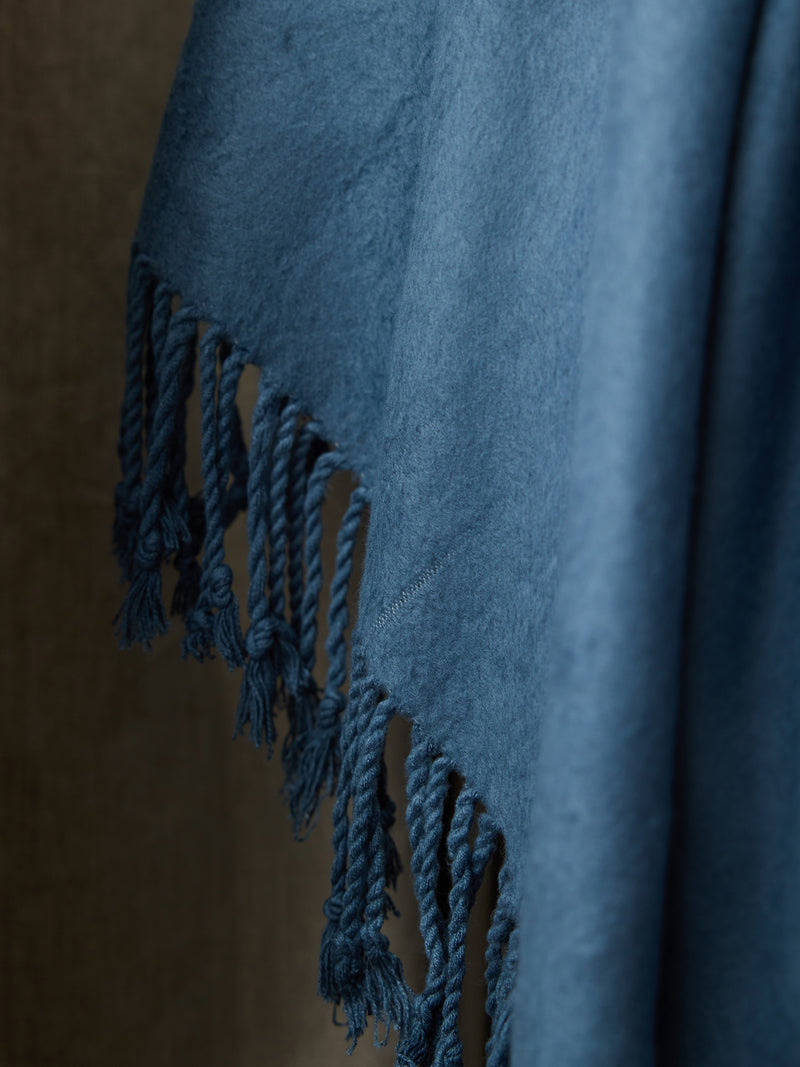 A close-up image of the Bamboo Tassel Throw by Cozy Earth in a soft blue hue. The fabric, appearing plush and fuzzy, is draped against a neutral background. Fringed tassel edges hang loosely, adding texture and detail to this luxurious throw.