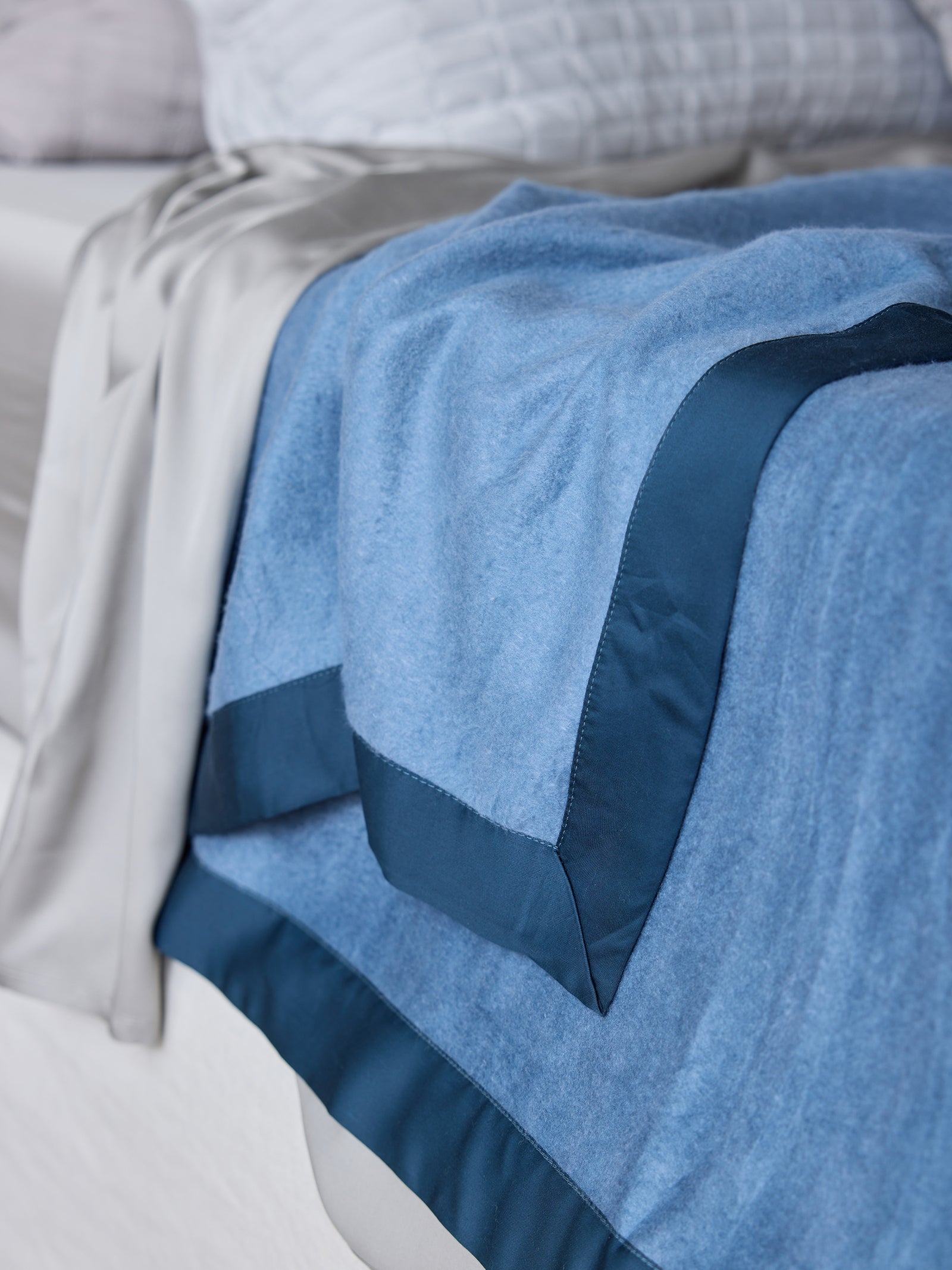 A close-up shot of a bed draped with Cozy Earth's Bamboo Blanket in soft blue, featuring a darker blue satin trim. Next to the blanket is a portion of light grey bedding, all set against a light grey bed frame. The background showcases white pillows with textured patterns. 