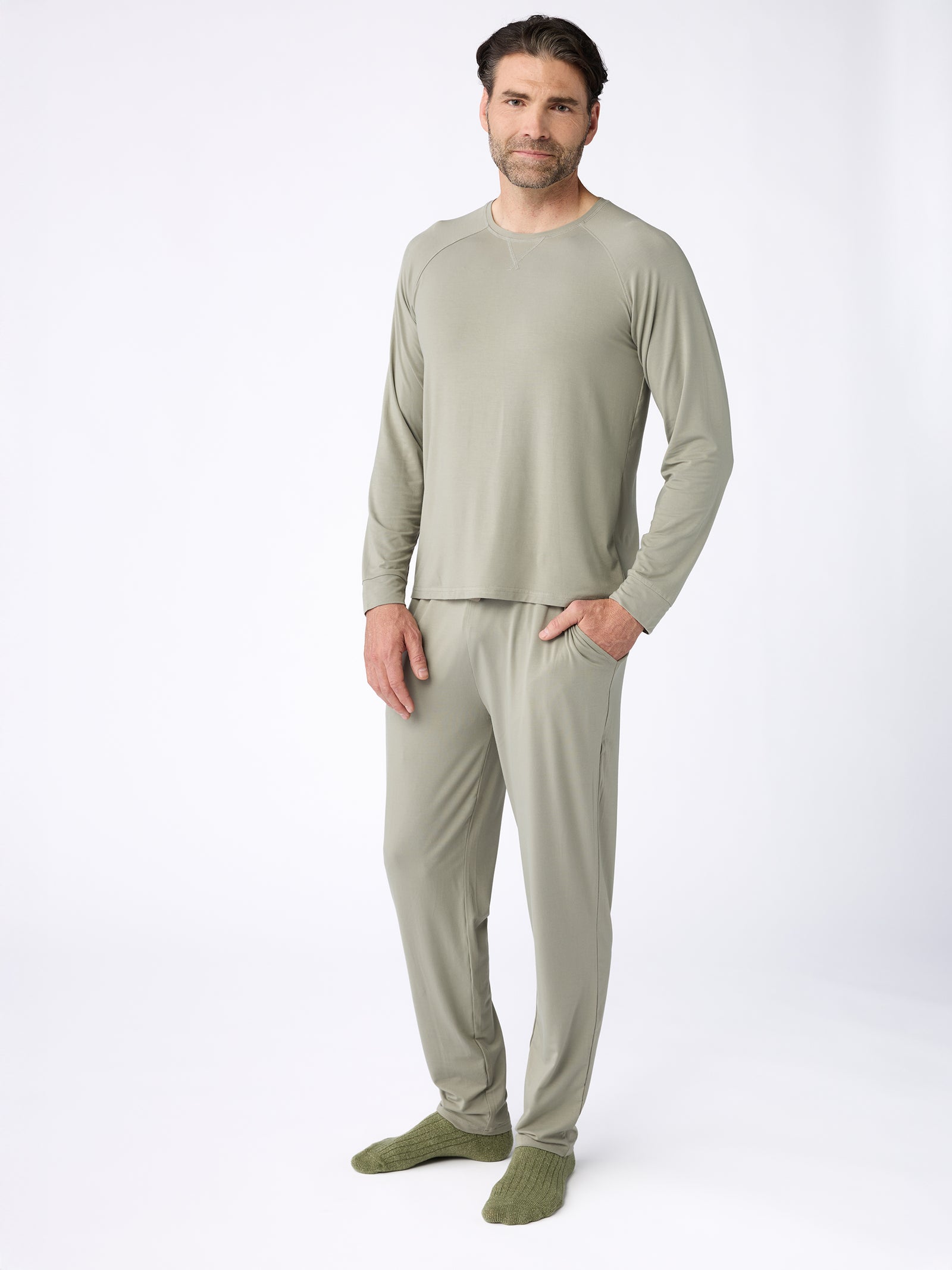 A person is standing against a plain background wearing matching light green Cozy Earth Men's Stretch-Knit Bamboo Long Sleeve pajamas and socks. Their hands are relaxed by their sides, and they have a neutral expression. The overall look is casual and comfortable. 