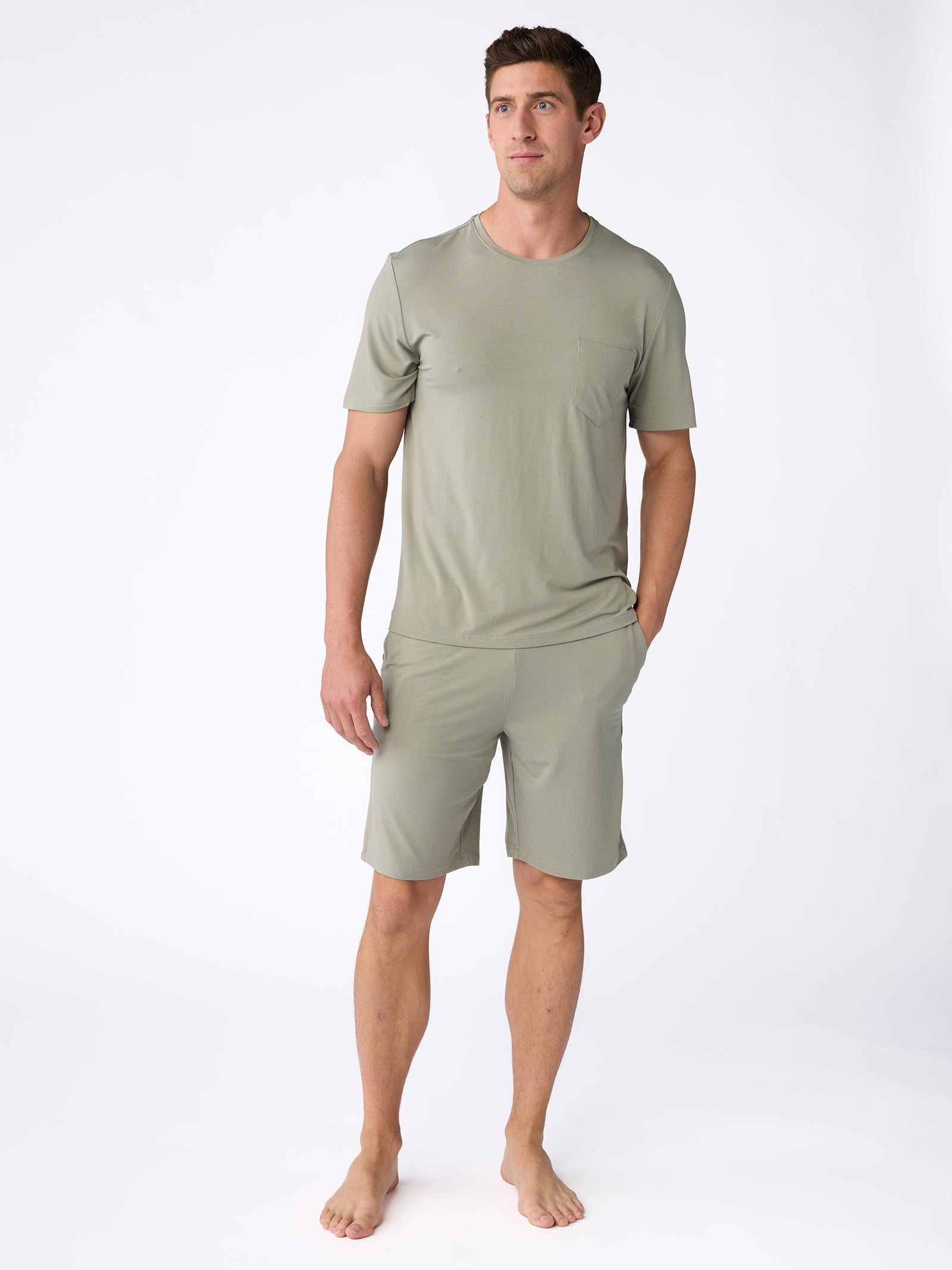 A man is standing barefoot against a white background, wearing an olive green short-sleeve shirt and matching Men's Stretch-Knit Bamboo Pajama Shorts by Cozy Earth. He has one hand in his pocket and is looking slightly to the side. 