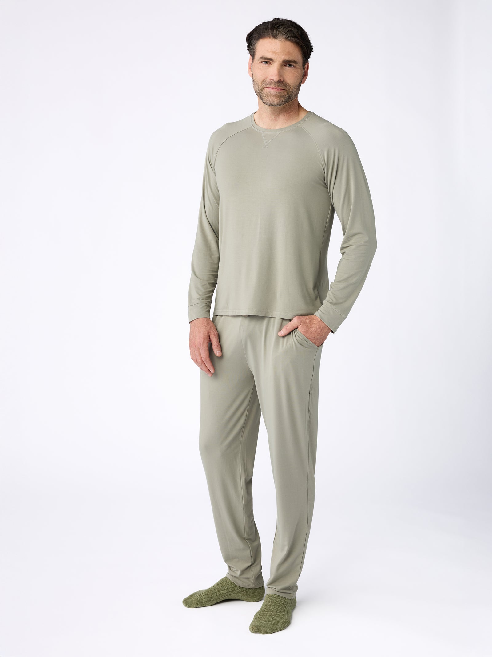 A man is standing against a plain white background, dressed in a light green long-sleeve pajama set from Cozy Earth. He pairs the Men's Stretch-Knit Bamboo Pajama Pants with matching socks, placing his hands in his pockets while maintaining a neutral expression. 