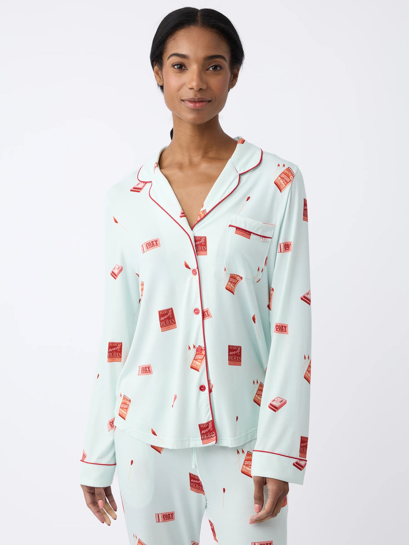 A woman models the Cozy Earth Women's Long Sleeve Bamboo Pajama Top in Stretch-Knit. The pajamas are light blue with red piping and feature a red book print. She stands against a plain white background, smiling slightly at the camera. 