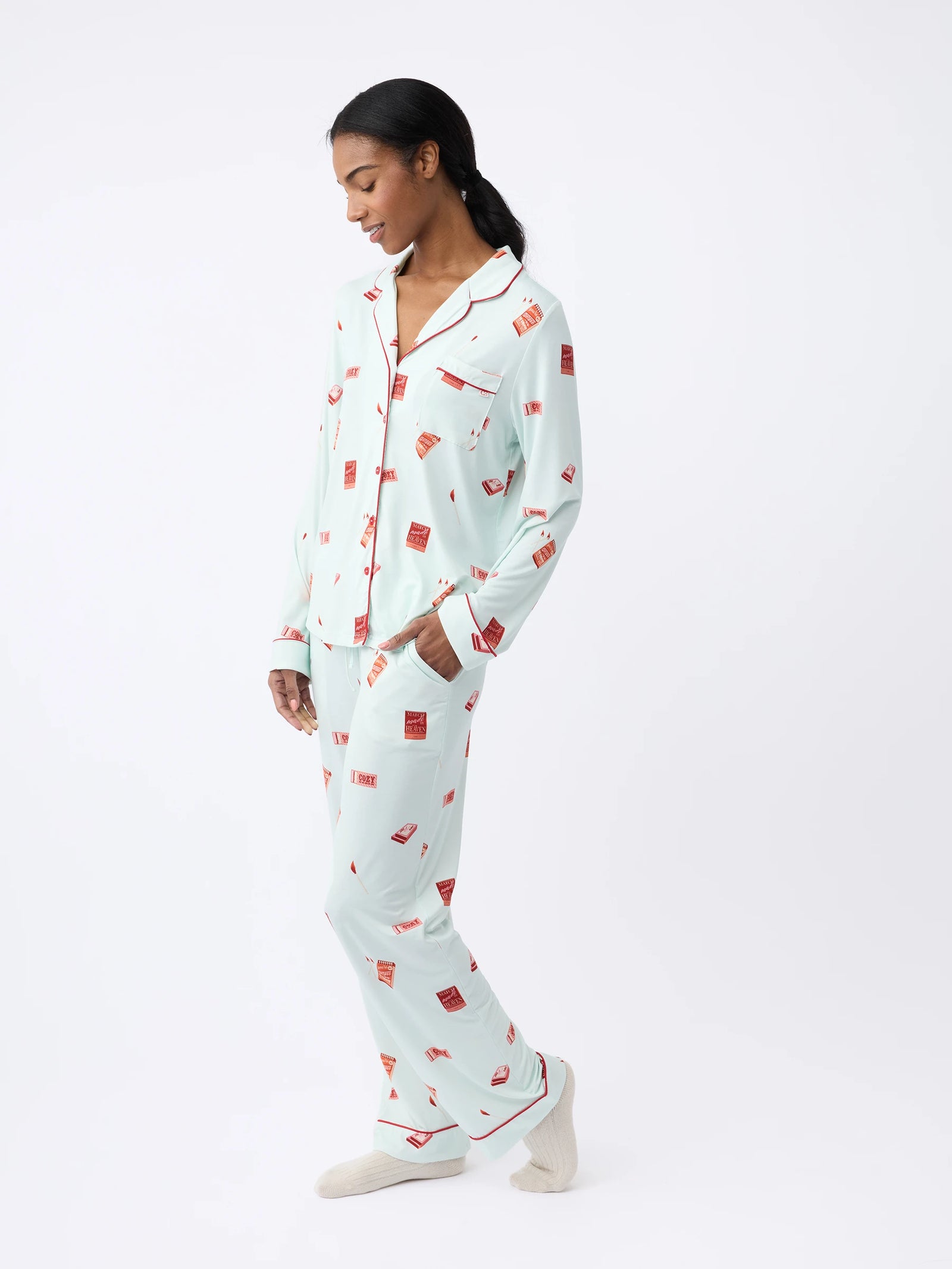 A woman wears Cozy Earth light blue Women's Stretch-Knit Bamboo Pajama Pant with red print and a matching long-sleeve button-up shirt, standing against a white background while looking down smiling slightly. 