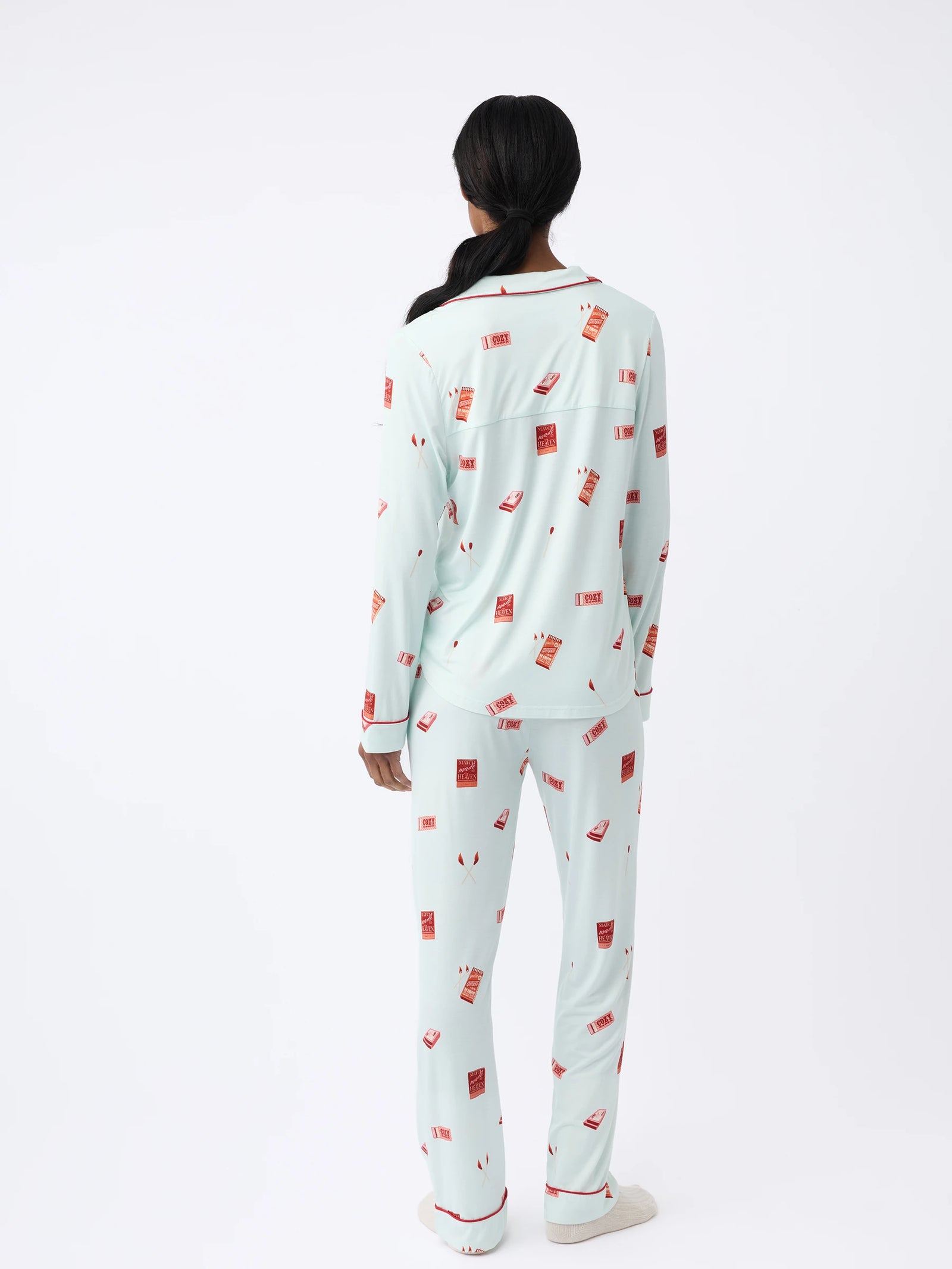 A woman with long dark hair, wearing a Cozy Earth Women's Stretch-Knit Long Sleeve Bamboo Pajama Set in light blue featuring a red and white print, stands back to the camera against a plain white background. 