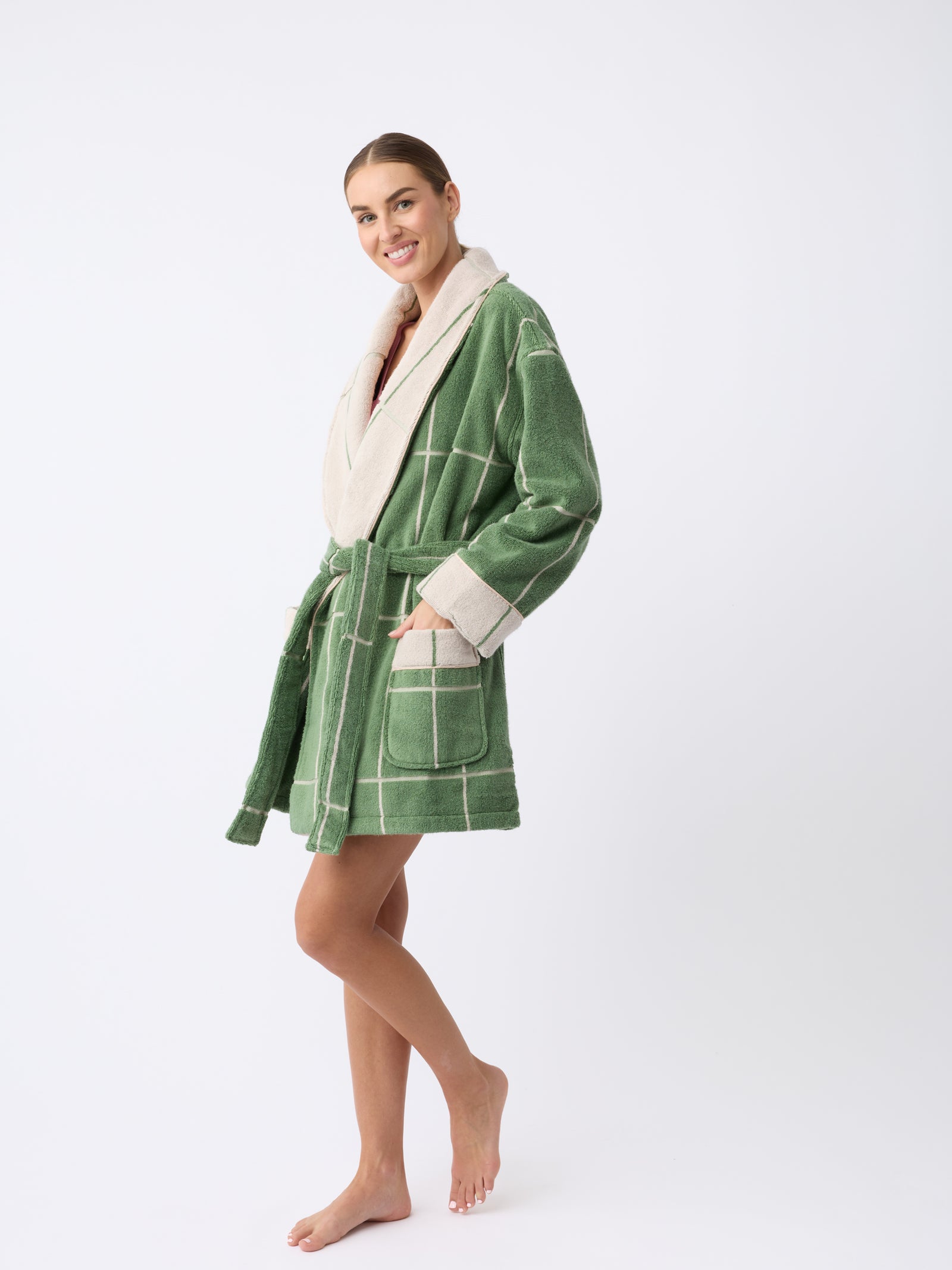 A smiling person stands barefoot in a Windowpane Resort Robe by Cozy Earth, featuring a green and white checkered pattern with cream-colored trim, posing against a plain white background. 