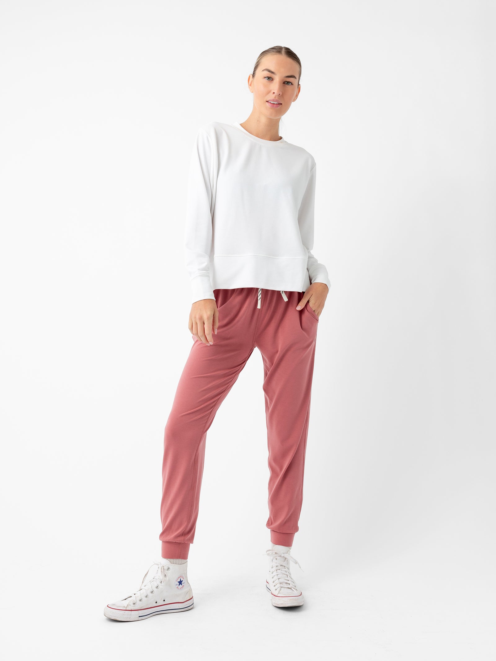 A person in a casual outfit stands against a plain white background, wearing a white long-sleeve top, the Cozy Earth Women's Studio Jogger in pink, and white sneakers with multicolored details. They stand with a relaxed posture, hands partially in pockets, and a slight smile. 