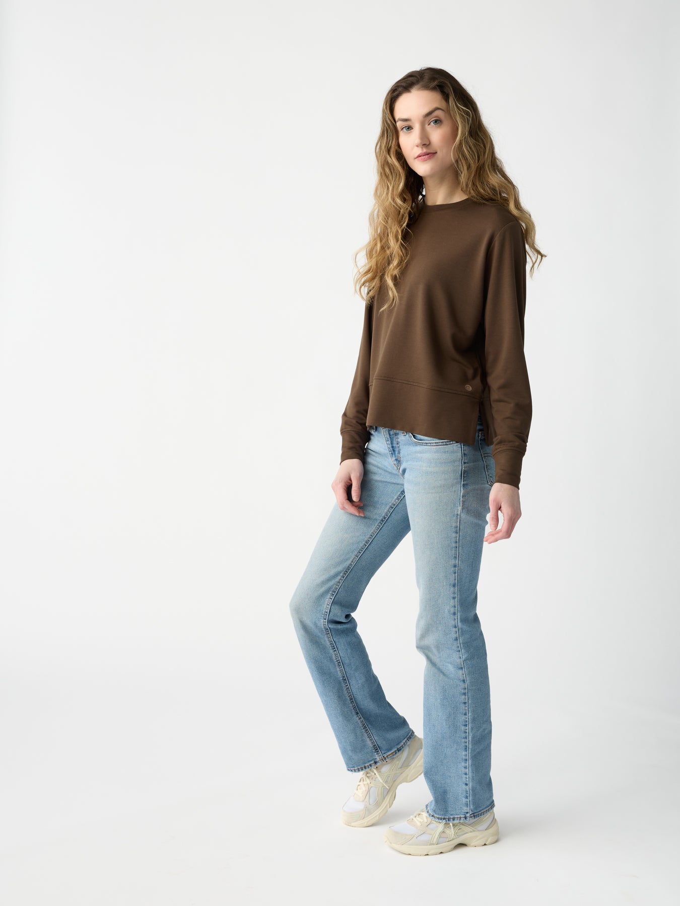 A person with long wavy hair wears a Cozy Earth Women's StudioLite Crewneck, light blue jeans, and white sneakers while standing against a plain white background. 