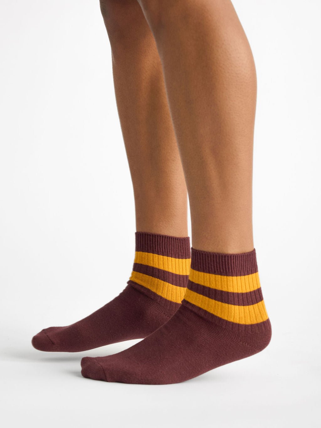 A person wearing Cozy Earth's Women's Sadie Socks in maroon with two horizontal mustard yellow stripes, covering the ankles, stands on a white surface. The image captures their legs from the knees down. 