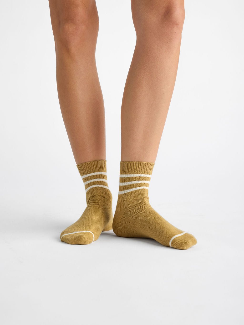 A person wearing Cozy Earth's Women's Sadie Sock in mustard yellow, featuring white stripes and accents at the toes and cuffs, stands on a white background, showcasing their lower legs and feet. 