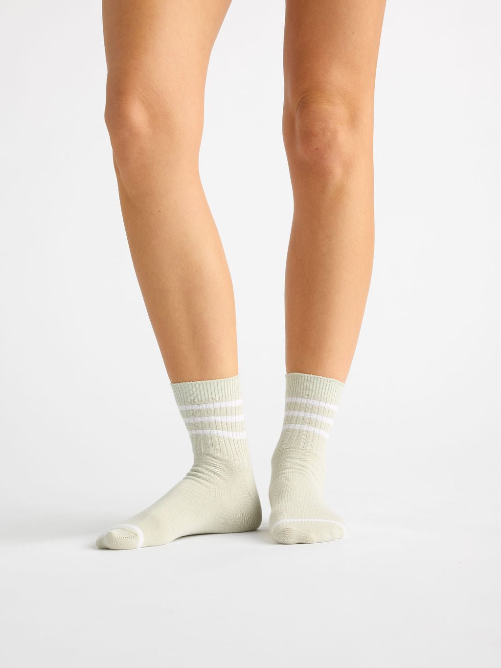 A person stands on a plain white background wearing Cozy Earth's Women's Sadie Sock in light pastel green with white stripes. The ribbed socks cover the ankles, and only their legs and feet are visible. 