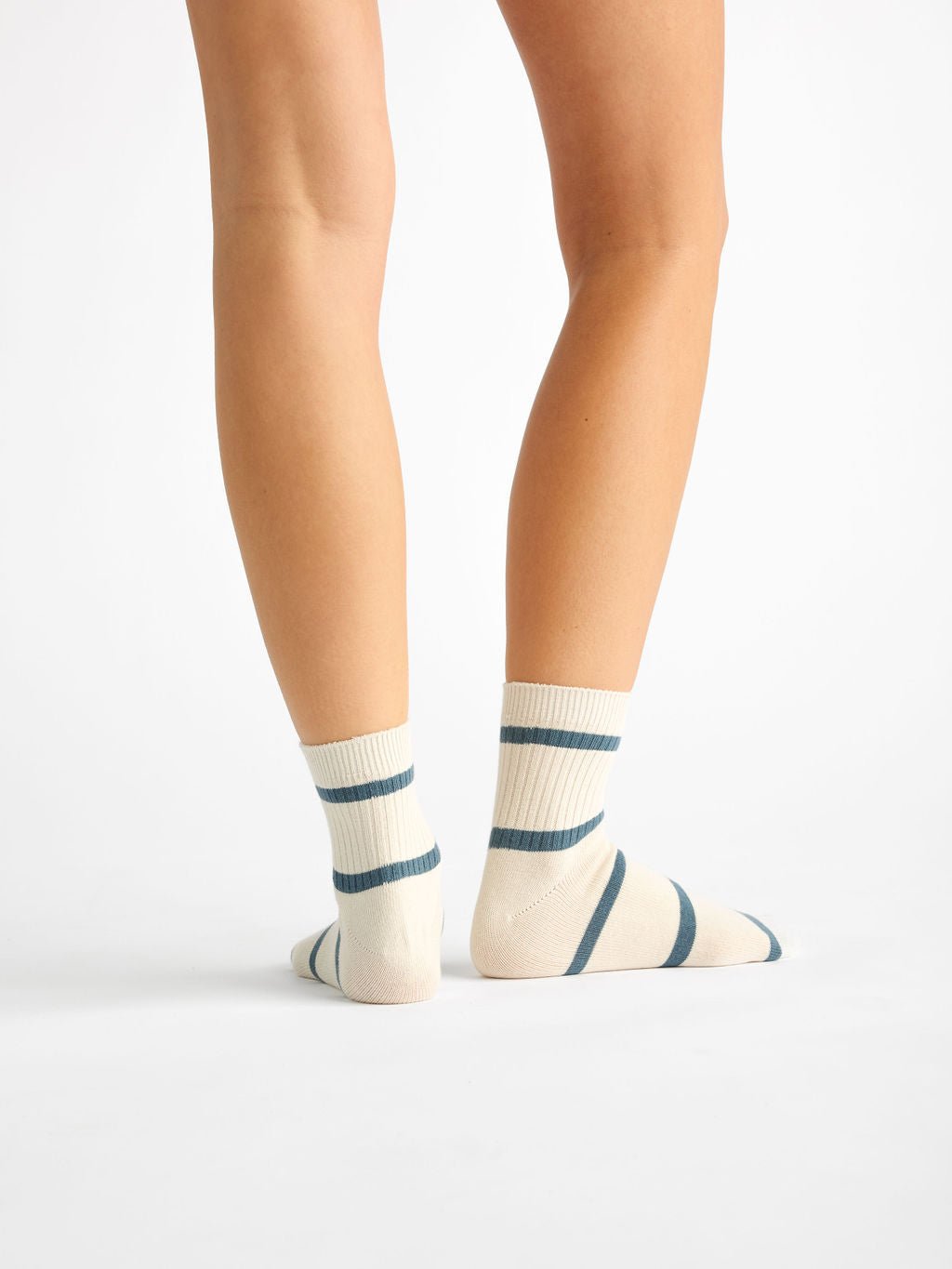 On a white background, a person wears Cozy Earth's Women's Sadie Sock, featuring cream with dark blue horizontal stripes. The image focuses on their lower legs and feet from the back. 