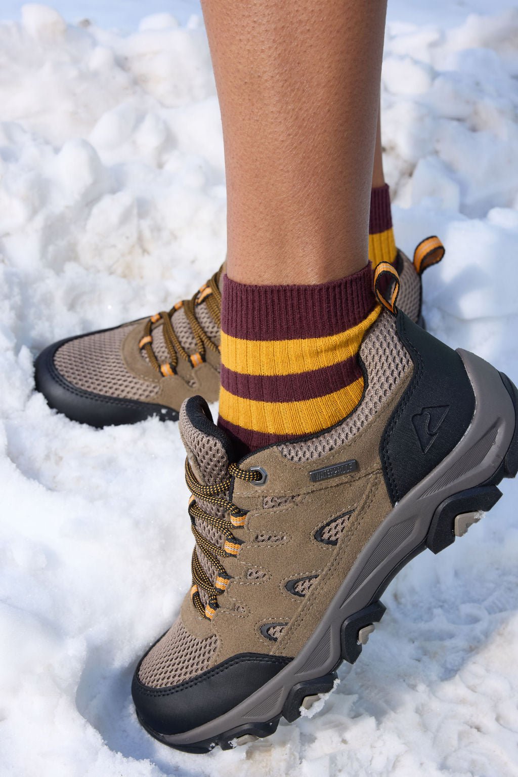 A person wearing Cozy Earth's Women's Sadie Sock in burgundy with two yellow ankle stripes, standing in snow wearing hiking boots