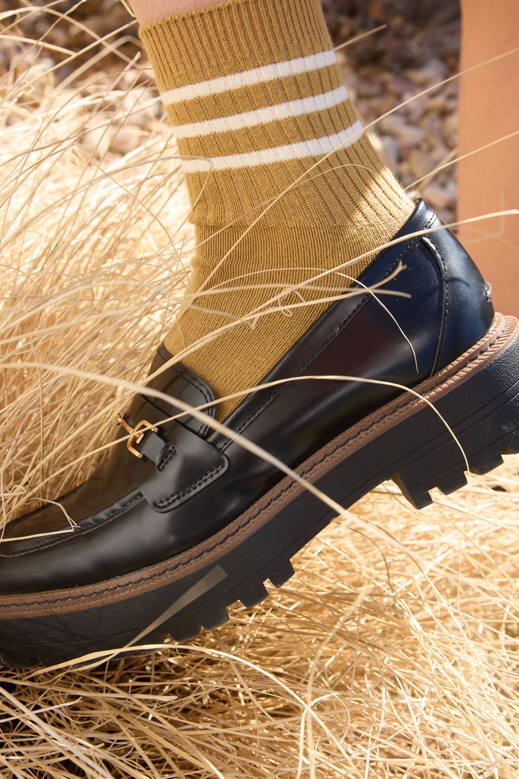 A person wearing Cozy Earth's Women's Sadie Sock in mustard yellow, featuring white stripes and accents wears black loafers and stands in a field of yellow grass. 