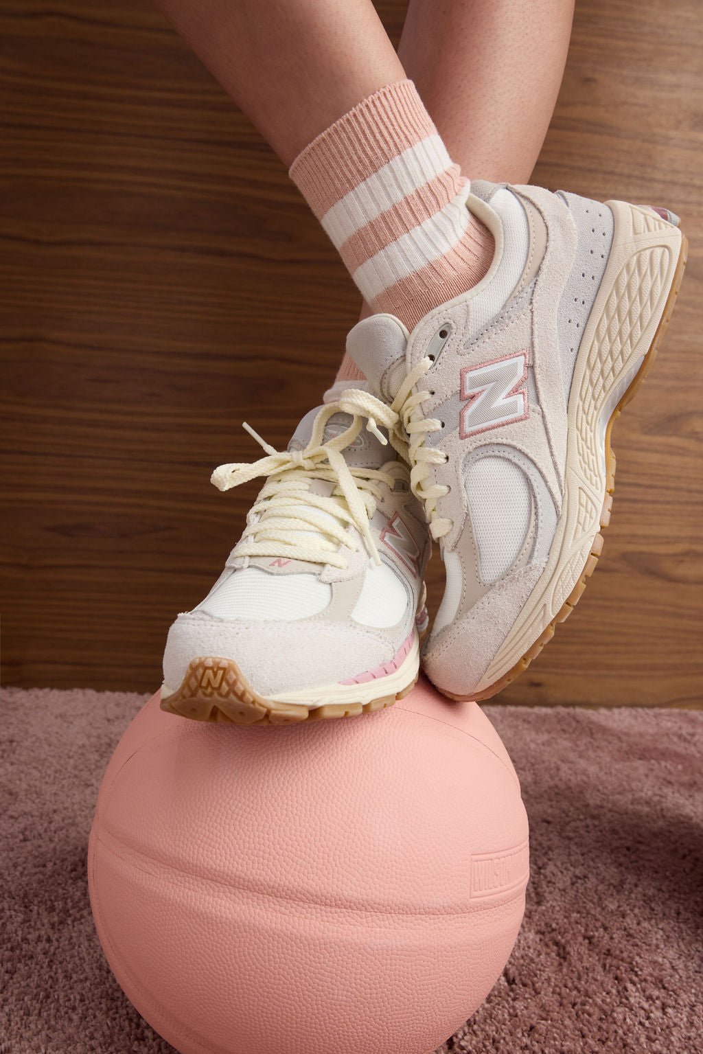 A person models the Women's Sadie Sock by Cozy Earth, featuring a peach shade with two white stripes, wearing sneakers and standing on a pink ball. 