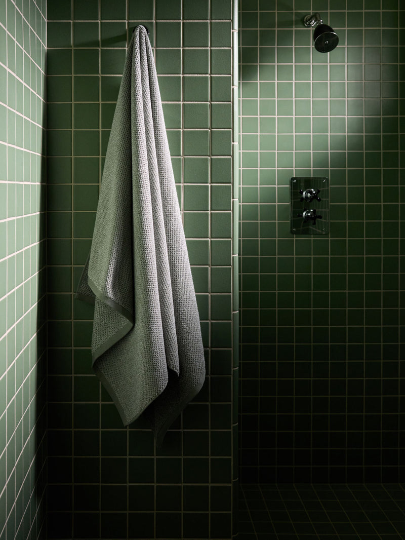 A Heathered Sage Nantucket Bath Sheet hangs in a green tiled shower.
