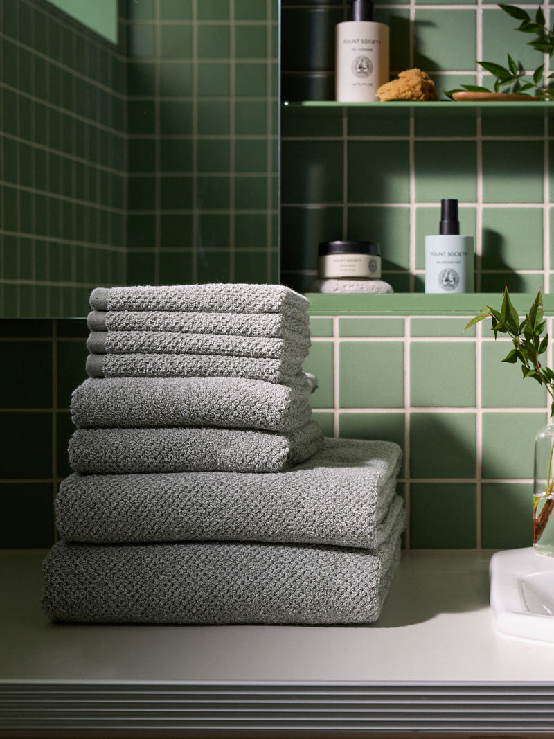 A stack of Nantucket towels and washcloths in Heathered Sage sit on a bathroom vanity countertop. There is a green tiled wall and shelves of skincare products in the background.