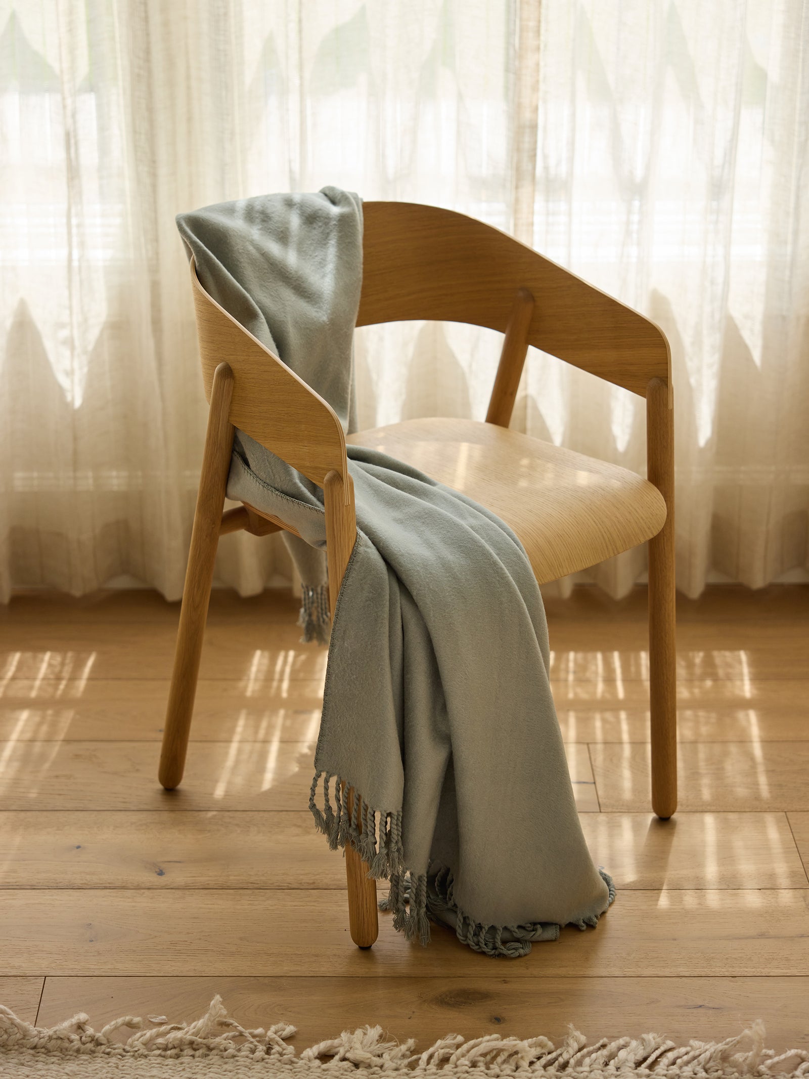 A wooden chair with a Cozy Earth Bamboo Tassel Throw in light blue draped over the backrest and seat sits in a sunlit room adorned with light-colored curtains and hardwood flooring. Natural light filters through the curtains, casting soft shadows on the floor. 