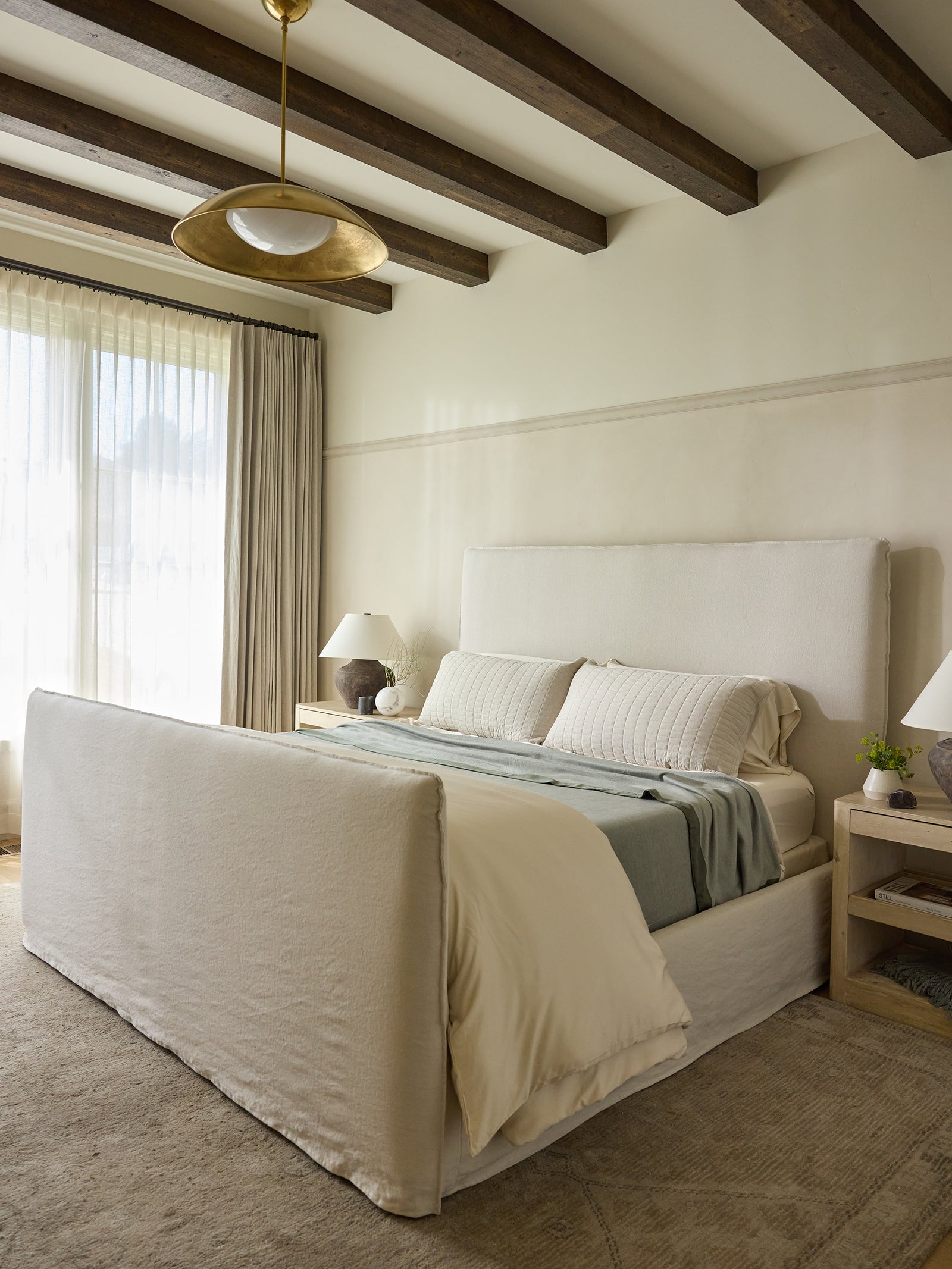 A cozy, modern bedroom features a large bed adorned with cozy Bamboo Blankets by Cozy Earth, in neutral tones along with matching pillows. The room boasts exposed wooden beams on the ceiling, a large window with sheer curtains letting in natural light, and side tables showcasing lamps and small potted plants. 