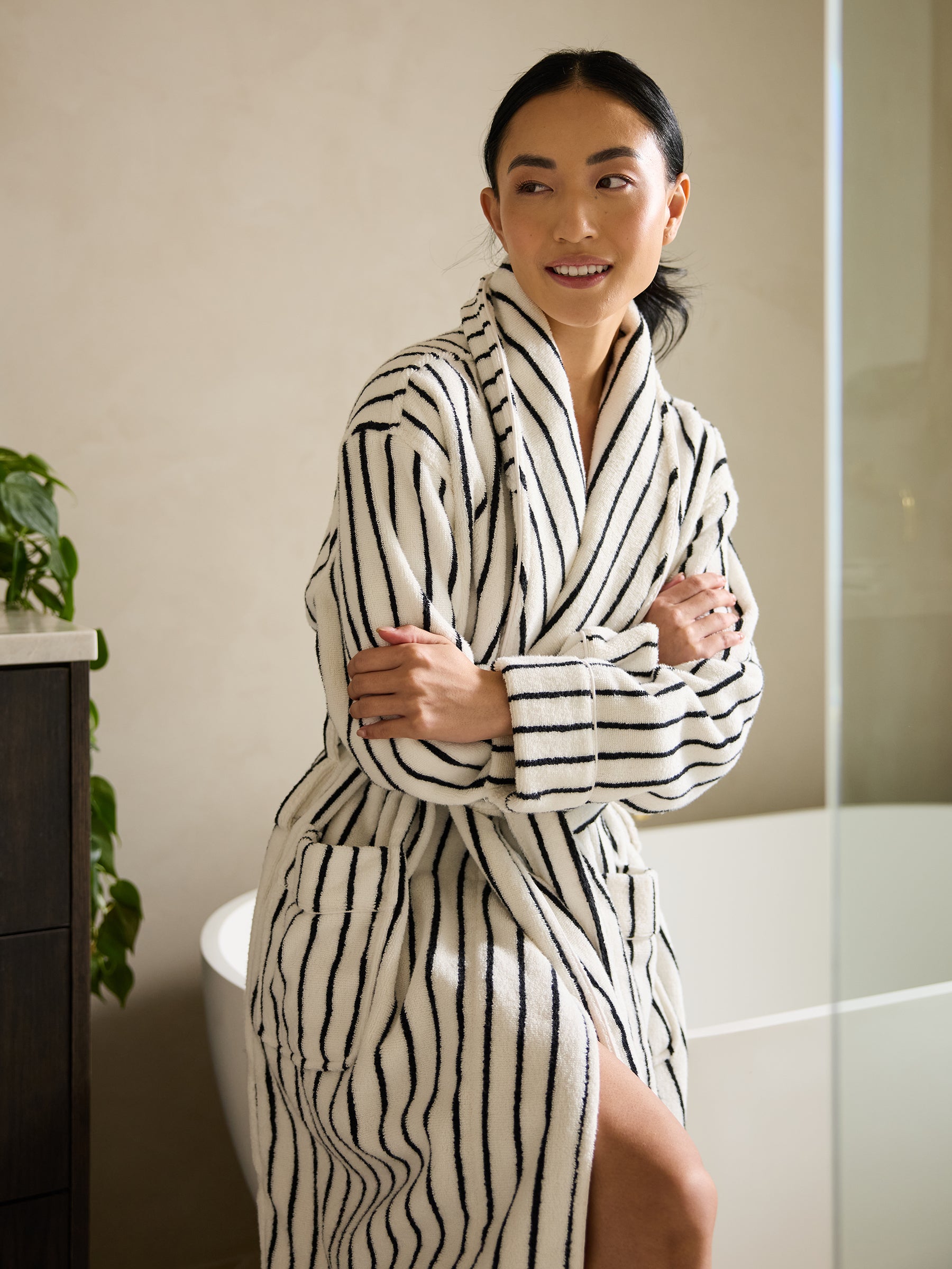 A person wearing the Luxe Bath Robe by Cozy Earth, featuring white fabric with black stripes, sits on the edge of a bathtub, smiling and looking to the side. A plant in the background complements the bathroom setting. |Color:Black Stripe