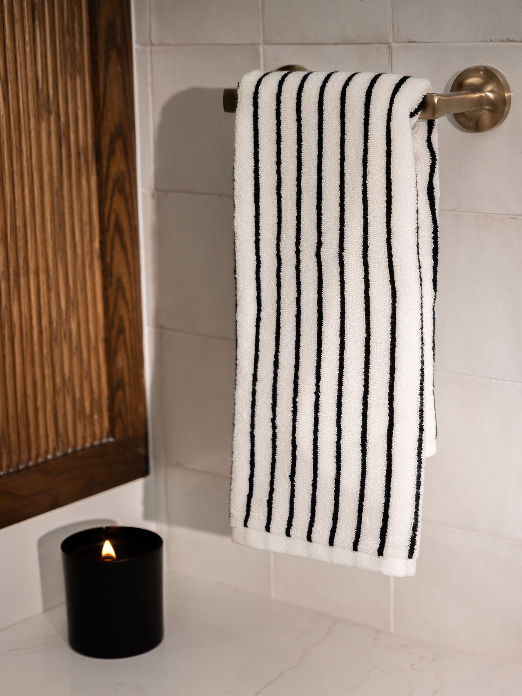 A neatly folded Luxe Hand Towel by Cozy Earth, featuring a white fabric with black vertical stripes, hangs on a silver towel rack mounted on a tiled wall. Below, a black candle container with a lit flame sits on a white countertop next to a wooden decor element. |Color:Black Stripe