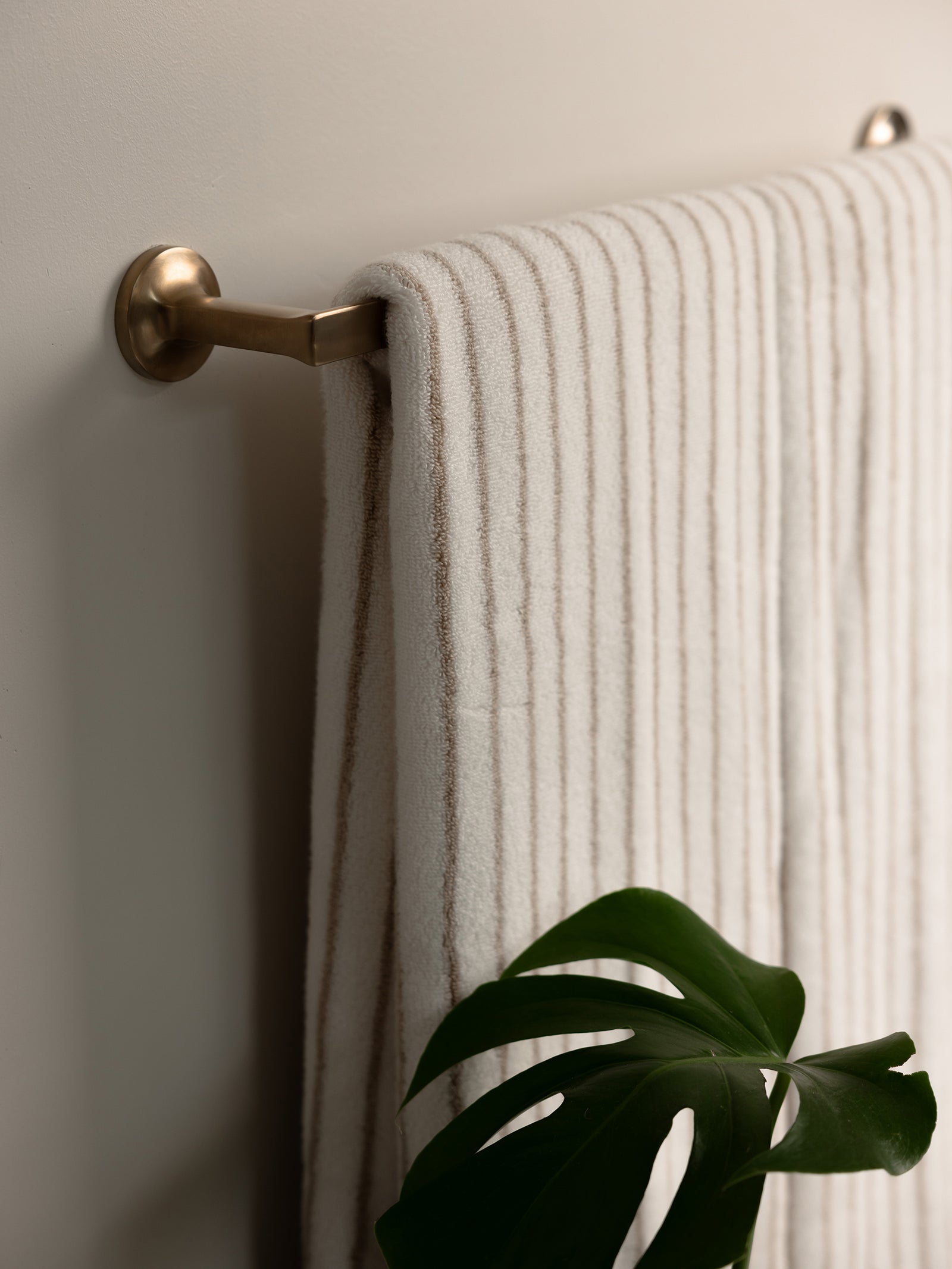 A neatly folded Luxe Bath Towel in textured beige from Cozy Earth hangs on a brass towel bar mounted on a light-colored wall. Below the towel, a green Monstera plant leaf is partially visible, adding a touch of nature to the clean and minimal bathroom setting. 