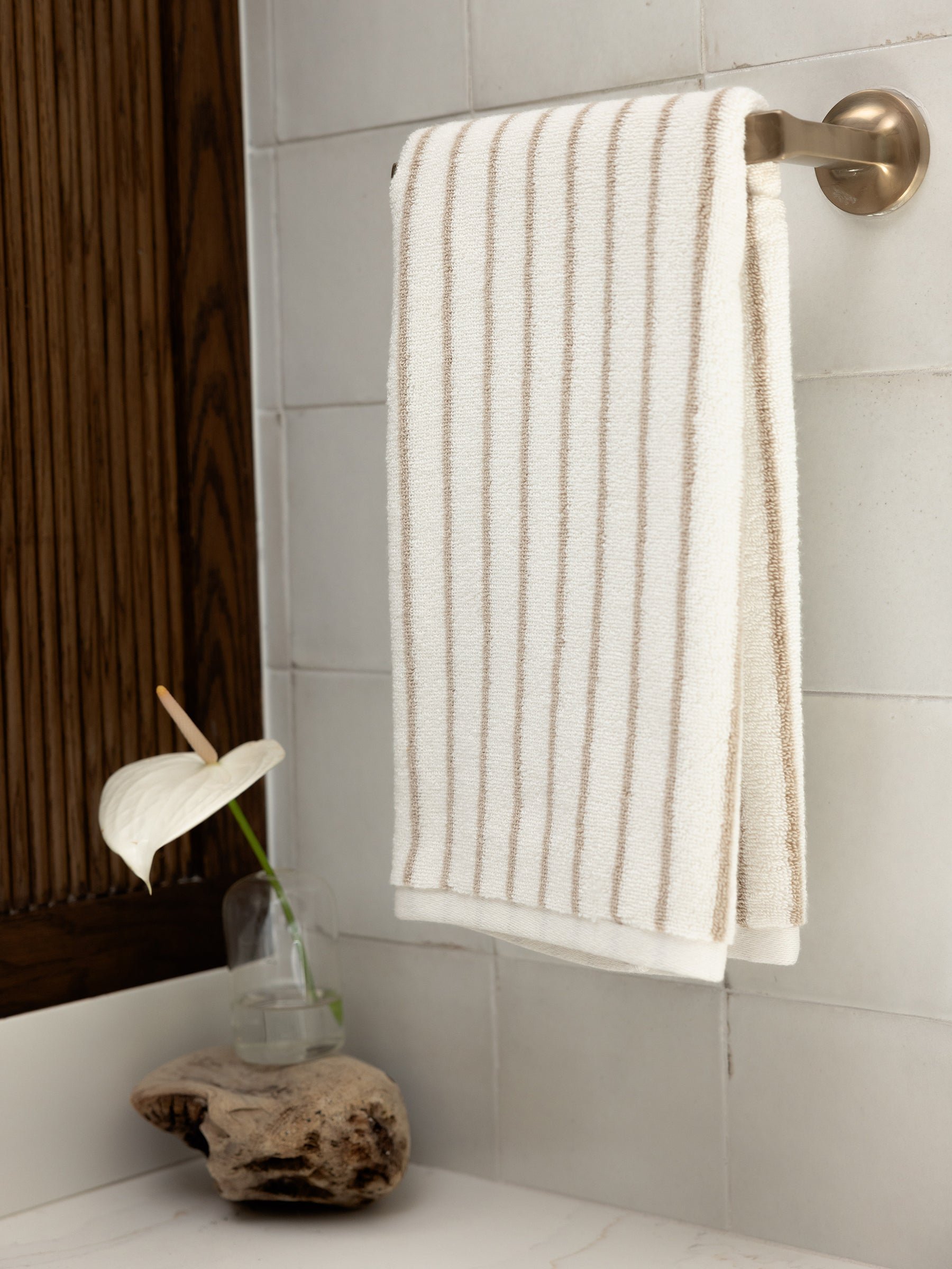 A neatly folded Luxe Hand Towel by Cozy Earth, featuring thin beige stripes, hangs on a silver towel bar in a bathroom. Below, a rustic wooden shelf holds a small glass vase with a single white anthurium flower. The bathroom walls are tiled with large, light-colored tiles. 