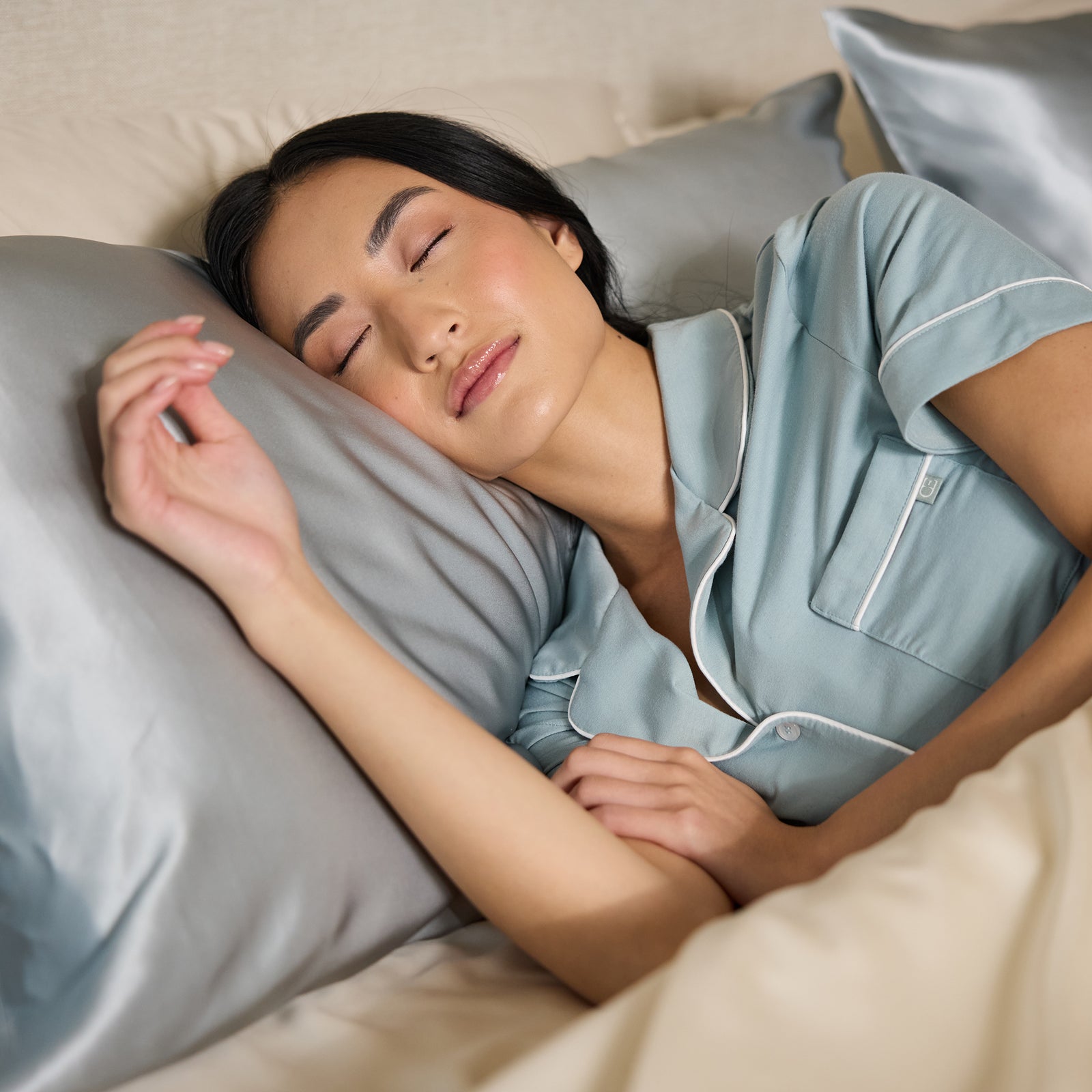 A person is sleeping peacefully on a bed, wearing blue pajamas, with their head resting on a silk pillowcase by Cozy Earth. The bedding is in a neutral color palette, exuding an aura of calmness and relaxation. 